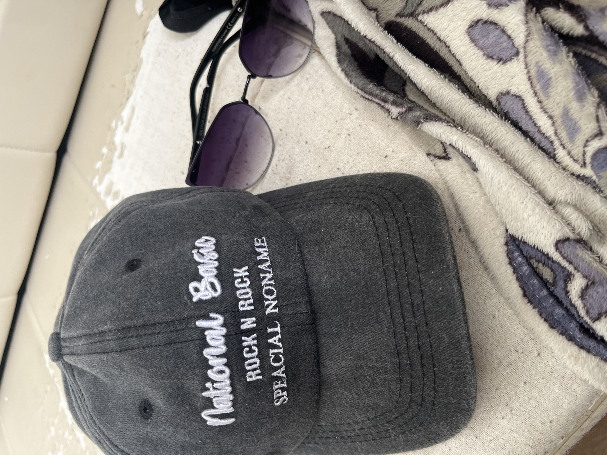 The image shows a close-up of a gray baseball cap with white lettering. The lettering on the cap reads "National Basic" in script font, and below that, "ROCK N ROCK" and "SPECiaL NONAME" in a block font. To the right of the cap, a pair of purple-tinted sunglasses with black frames is visible. The objects are placed on a textured, light-colored surface, possibly a towel or blanket, with a pattern of gray and purple shapes. The background is out of focus but appears to be a light-colored, upholstered surface, possibly a couch or seating area. There are no people or identifiable landmarks from Chelyabinsk, Russia in the image.