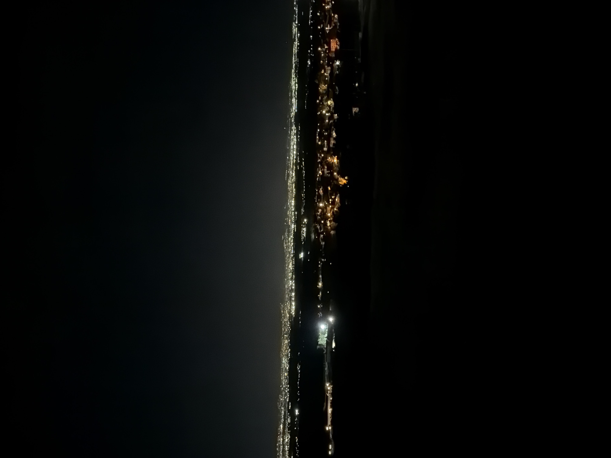 This is a nighttime, wide-angle shot of a city at dusk or night. The foreground is dominated by darkness, with a slight elevation suggesting the viewpoint is from a hill. In the mid-ground, lights from a town or city are visible, with a dense cluster of yellow and white lights indicating buildings and streets. Some lights appear to be brighter, possibly from streetlights or larger structures. The horizon line separates the illuminated city from a dark, clear sky. The overall impression is of a sprawling urban area seen from a distance at night. The scene is identified as being in Voynegovtsi, Bulgaria. There are no people or distinct objects in the foreground. The lighting suggests artificial illumination from the urban area.