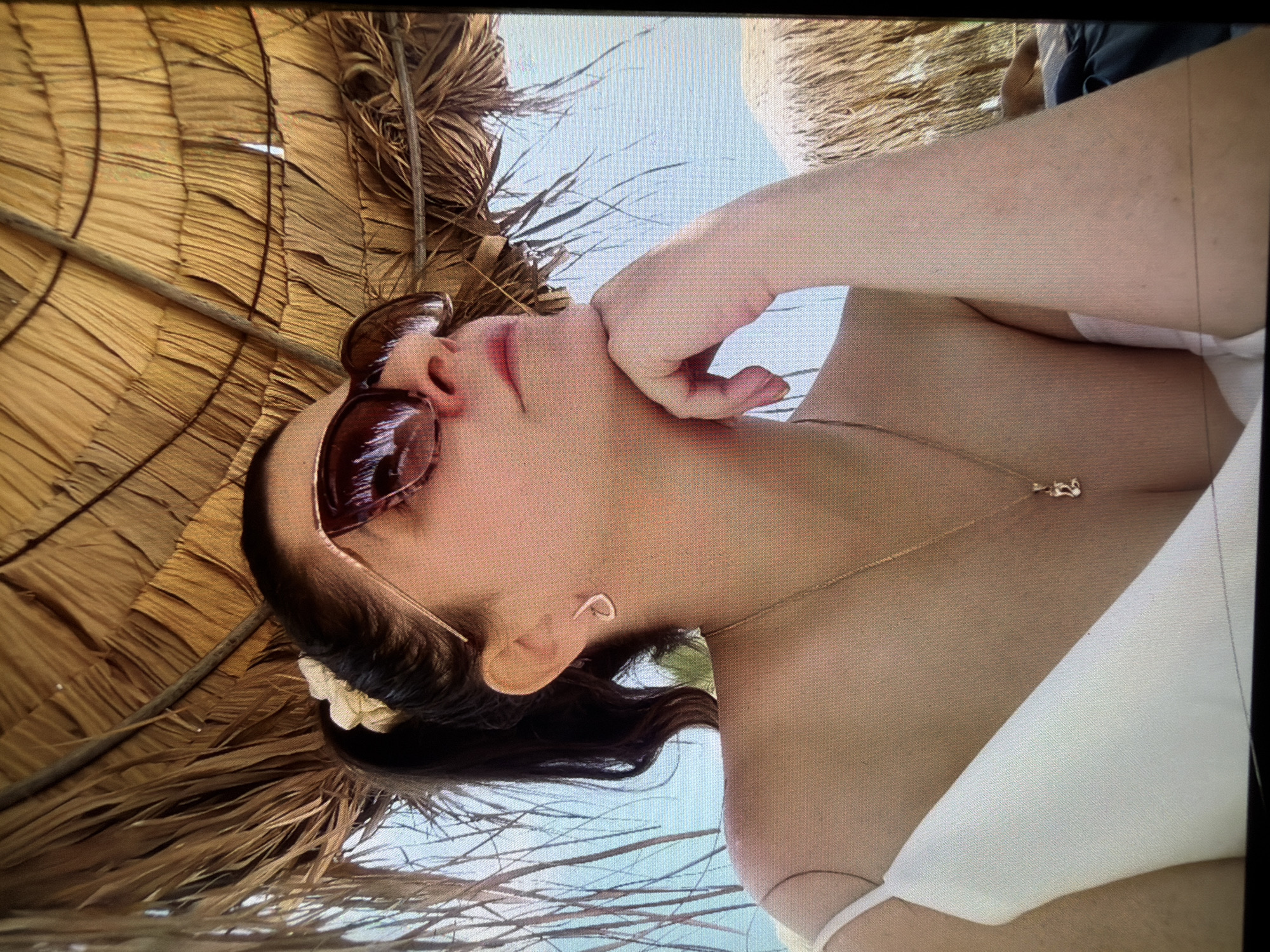 A woman wearing sunglasses and a white dress is sitting under a thatched roof at the beach. She is looking up and to the side, with her chin resting on her hand. A necklace with a pendant is visible on her chest. The background shows the sky and the ocean, along with another thatched roof structure.