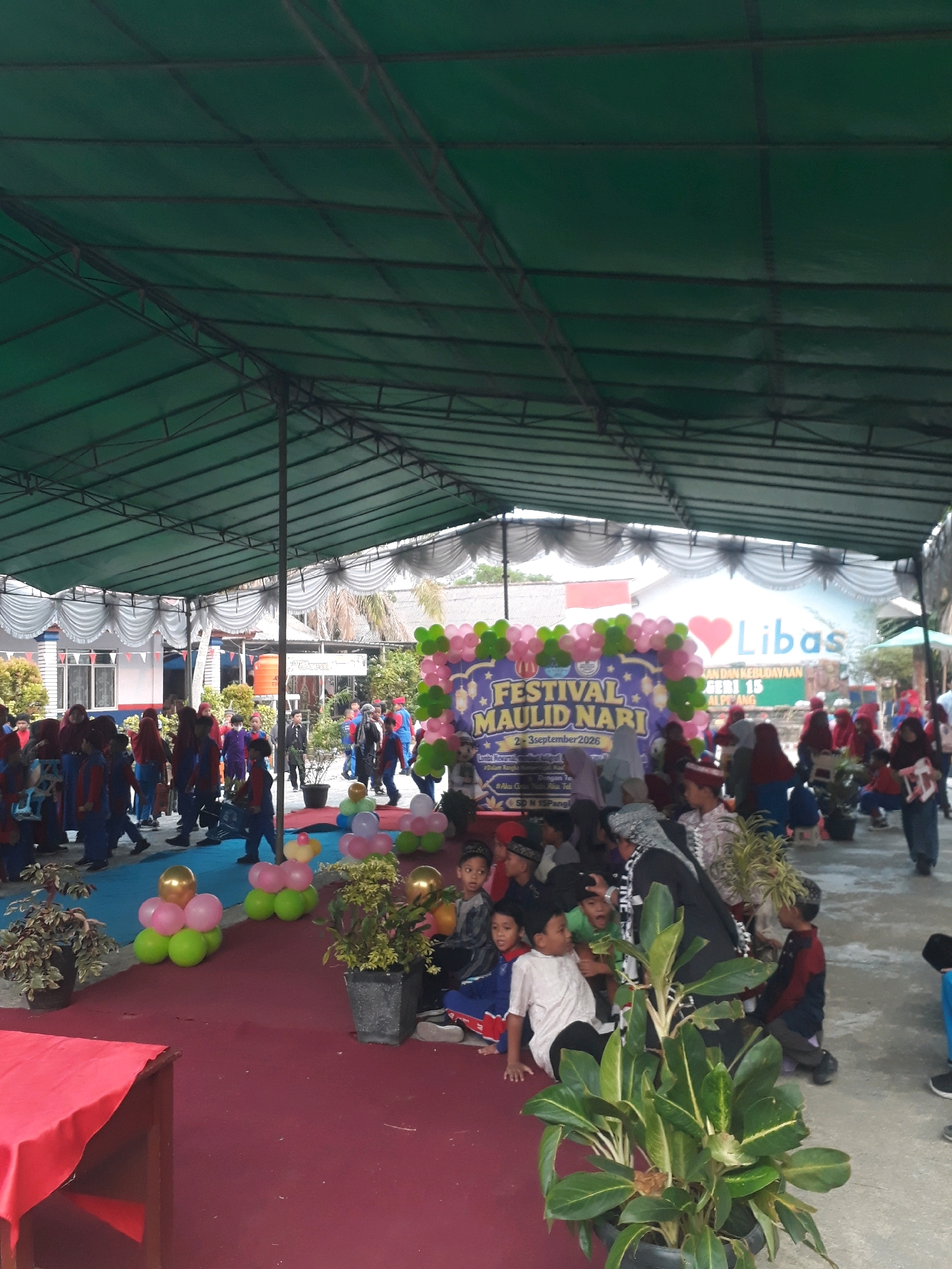 This outdoor event appears to be taking place under a large green tarp supported by metal poles, providing shade for a gathering of people. The ground is covered with a reddish carpet. In the background, there is a building with windows and some trees. A prominent banner on the right side of the image reads "FESTIVAL MAULID NARI" and "2-3September 2026". There are also several balloons, including some in the shape of animals, decorating the area. Many children are present, some wearing uniforms and traditional headwear. Some adults are also visible, supervising or participating in the event. The overall atmosphere suggests a festive community celebration. Based on the banner mentioning "SD N 15 Pangkalpinang" and "Telp.", it is highly probable that this event is located at SD Negeri 15 Pangkalpinang in Pangkalpinang, Indonesia.