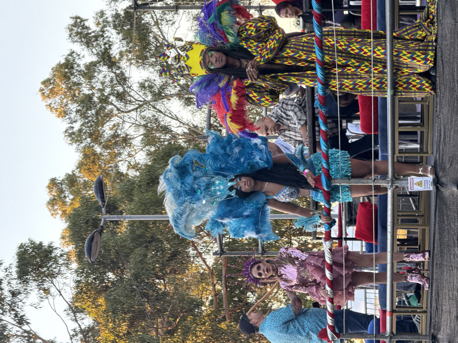 An outdoor scene in Cape Town, South Africa, depicts an event on an elevated, multi-tiered platform, illuminated by late afternoon sunlight. Three individuals in elaborate costumes are positioned in the foreground.
The leftmost individual is a fair-skinned female with dark hair, wearing a long-sleeved, shimmering rose-gold dress with ruffled tiers, a spiky purple headpiece, and metallic pink high-heeled boots. Her hands are positioned in front of her waist.
The central figure is a dark-skinned female with long dark hair, adorned with a large, intricate blue feathered and sequined headpiece, a matching blue sequined bra top, and a short, fringed skirt. She wears white open-toed sandals, and her right hand rests on a red and blue striped railing.
The rightmost individual, dark-skinned with dreadlocks, wears a long robe patterned with yellow, green, red, and black geometric shapes. They are crowned with an elaborate yellow headpiece featuring colorful feathers and possess additional large, multi-colored feathered shoulder pieces. Their head is slightly bowed, and hands are clasped at waist level.
In the mid-ground, behind the costumed figures, several other individuals are present on the platform. To the far left, a man in a light blue patterned shirt and dark cap faces away from the camera. Behind the central figure, a balding man in a plaid shirt is seated, looking right. To the far right, a woman with dark hair in a dark top is partially visible. The platform features red cushioned seating, and a white warning sign with "DANGER" in black text and a yellow triangle is visible on the floor. The background consists of tall green trees under a clear, pale sky. A streetlamp structure with two dark fixtures is visible above the trees.
