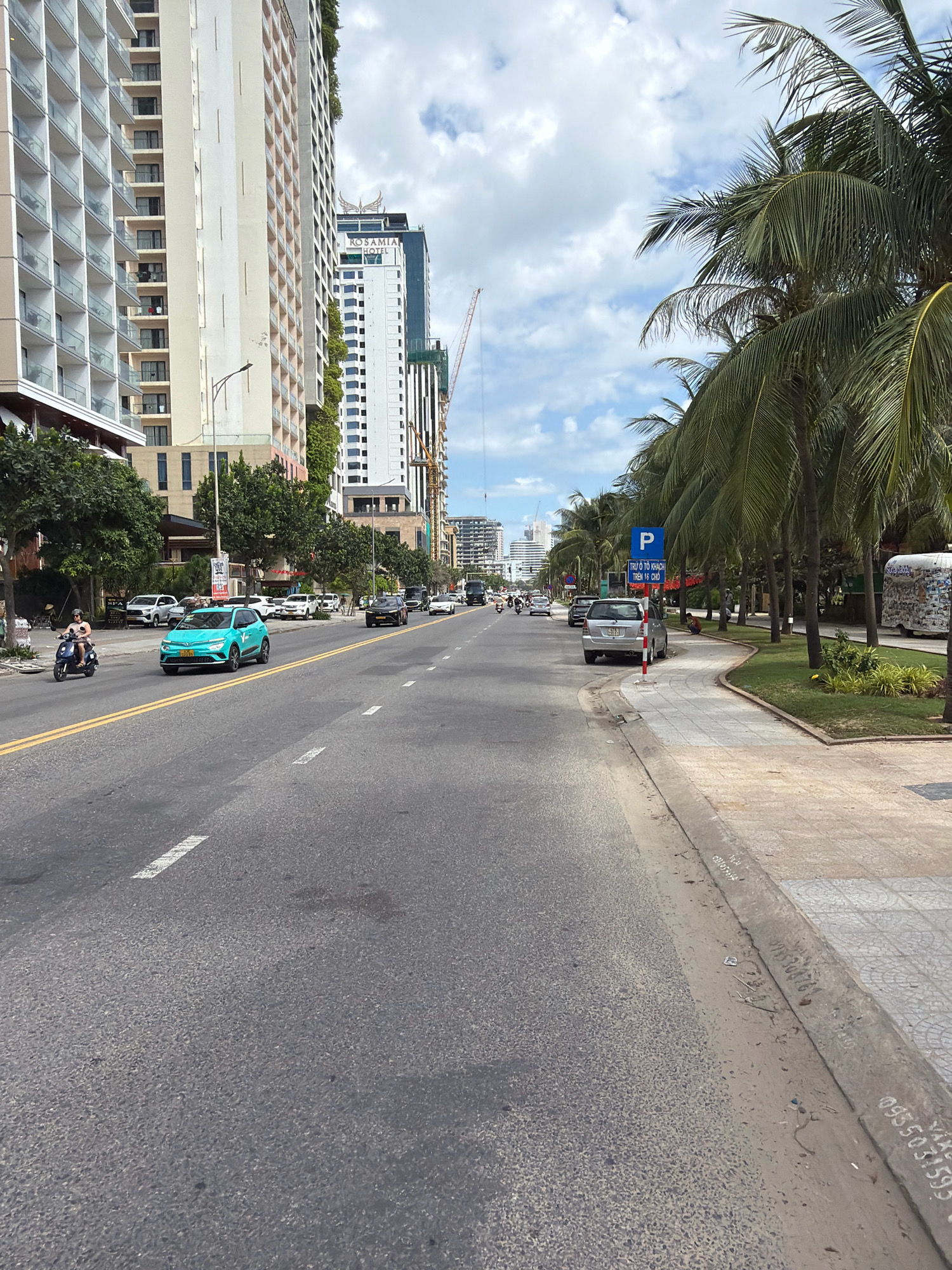 The image depicts a street scene in the city of Đà Nẵng, Vietnam, likely along a coastal road. The perspective is from the middle of a paved road with two lanes in each direction, separated by yellow lines. On the left side of the road, tall, modern buildings with multiple balconies are visible. Green trees line the sidewalk in front of these buildings. A person is riding a blue scooter in the left lane, moving away from the camera. Several cars are also on the road, including a teal-colored hatchback in the foreground and a silver SUV on the right side of the road.
The right side of the road is characterized by a row of palm trees and a sidewalk. A blue parking sign with Vietnamese text is posted. Further down the sidewalk, a silver car is parked. The sky is partly cloudy. The overall impression is of a busy urban environment with tropical vegetation and modern architecture. The image includes various elements of urban infrastructure and traffic.