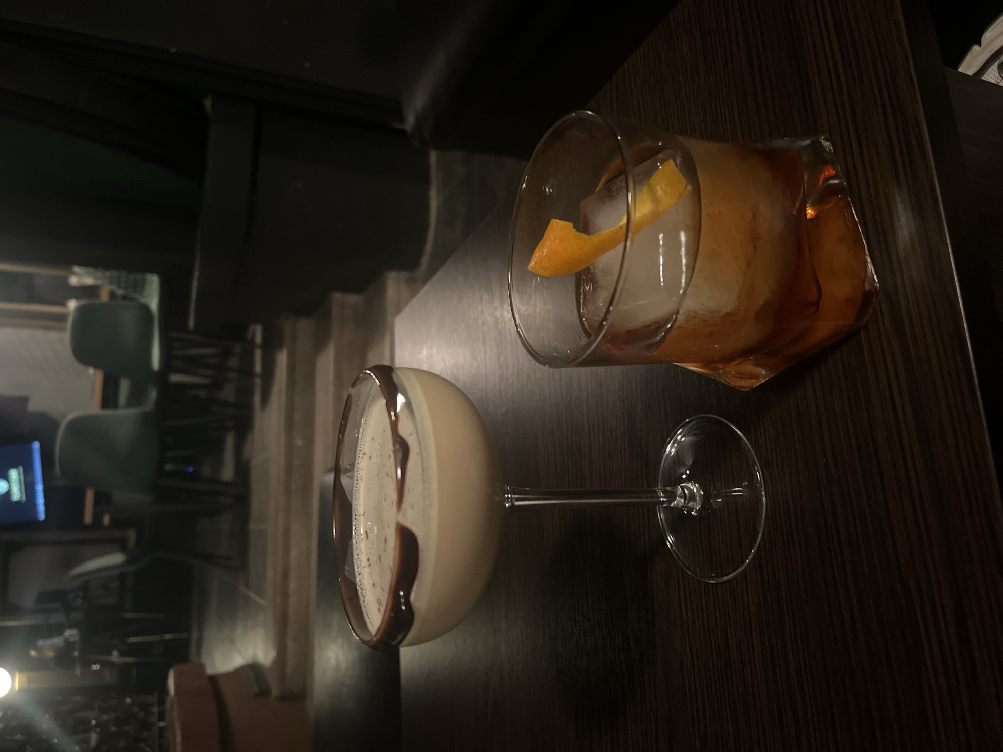 This image depicts an indoor establishment, identified as being in Varna, Bulgaria, likely a bar or lounge, characterized by low lighting and dark furnishings. In the foreground, positioned on a dark brown, wood-grain table, are two alcoholic beverages. To the left, a light-beige cocktail with a frothy top is presented in a clear, stemmed coupe glass, its rim adorned with a dark chocolate drizzle. To the right, a second cocktail, dark amber in color, is served in a clear, irregularly shaped lowball glass, containing a large square ice cube and garnished with an orange peel.
The background, heavily blurred and dimly lit, reveals a tiered seating arrangement. Multiple dark-toned chairs with light-green or teal upholstery are visible, some pushed under tables. A television screen with a visible logo or text, partially decipherable as "TURKISH," is present in the upper-right portion of the background. Dark wall panels or furniture elements complete the subdued interior setting. No individuals are discernible within the frame.