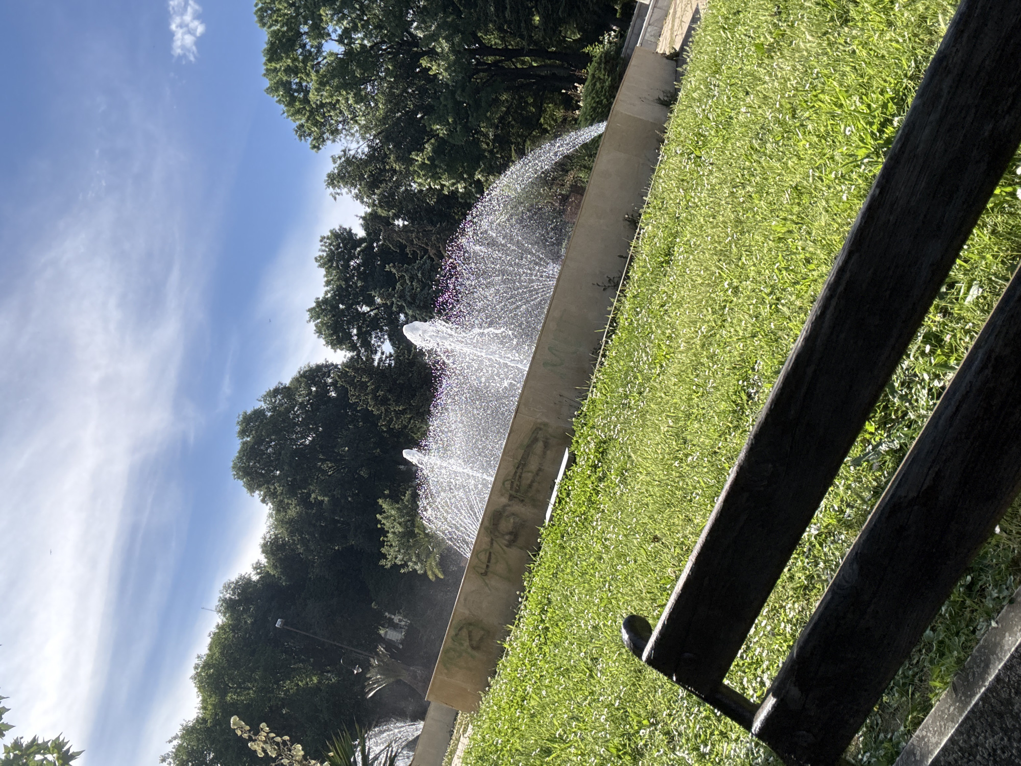 The image shows a park scene with a fountain spraying water in the foreground. The fountain has a modern, minimalist design with a concrete basin. Lush green grass covers the ground around the fountain, dotted with small white flowers. In the background, mature trees with dense foliage create a natural backdrop. A portion of a dark wooden bench is visible in the lower right corner, suggesting a place for relaxation or observation.
The activity taking place is the fountain's operation, with water arcing and scattering in the sunlight, creating a dynamic visual effect. The bright sunlight and clear blue sky, with wispy white clouds, indicate it is daytime, likely late morning or afternoon. The overall atmosphere appears peaceful and serene.
There is no visible text in the image. The location cues, such as the style of the fountain and the type of trees, along with the provided location context of Varna, Bulgaria, help to place the scene. The image evokes a sense of calm and appreciation for nature and public spaces.