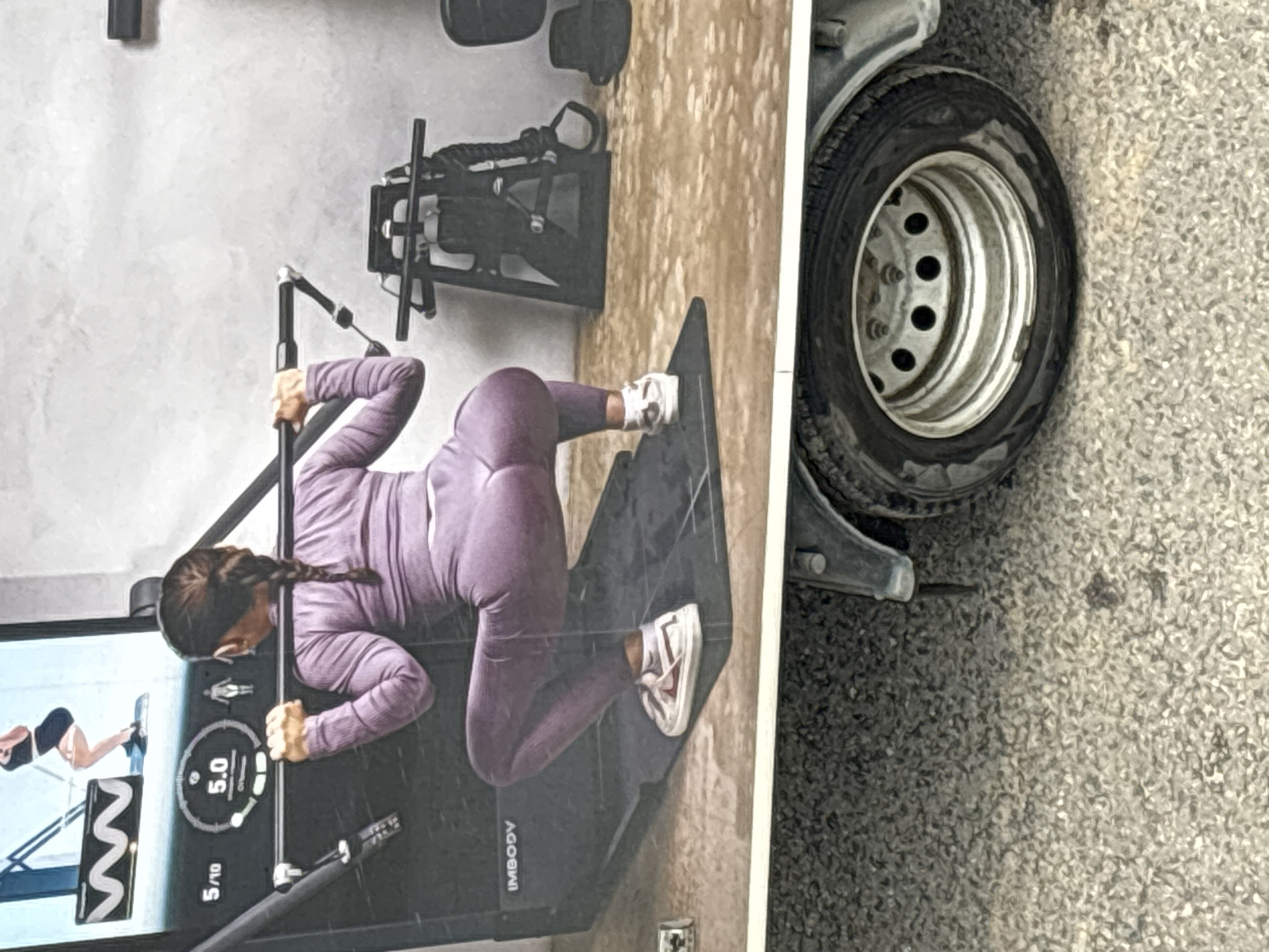The image shows a close-up view of a mural on the side of a vehicle, possibly a truck or trailer, parked outdoors. The mural depicts a woman in athletic wear performing a squat exercise. She is holding a barbell across her shoulders and is wearing a purple athletic outfit and white sneakers. Behind her, a digital display on what appears to be a fitness machine shows "5.0" and "5/10". Additional gym equipment is also visible in the mural. In the foreground, the lower part of the vehicle is visible, including a wheel with a silver rim and black tire, parked on a surface covered in gravel. The mural is positioned above this vehicle. There is no indication of the specific location within Ostrov, Russia, being identifiable from the image itself; the context of Ostrov, Russia, is provided externally.