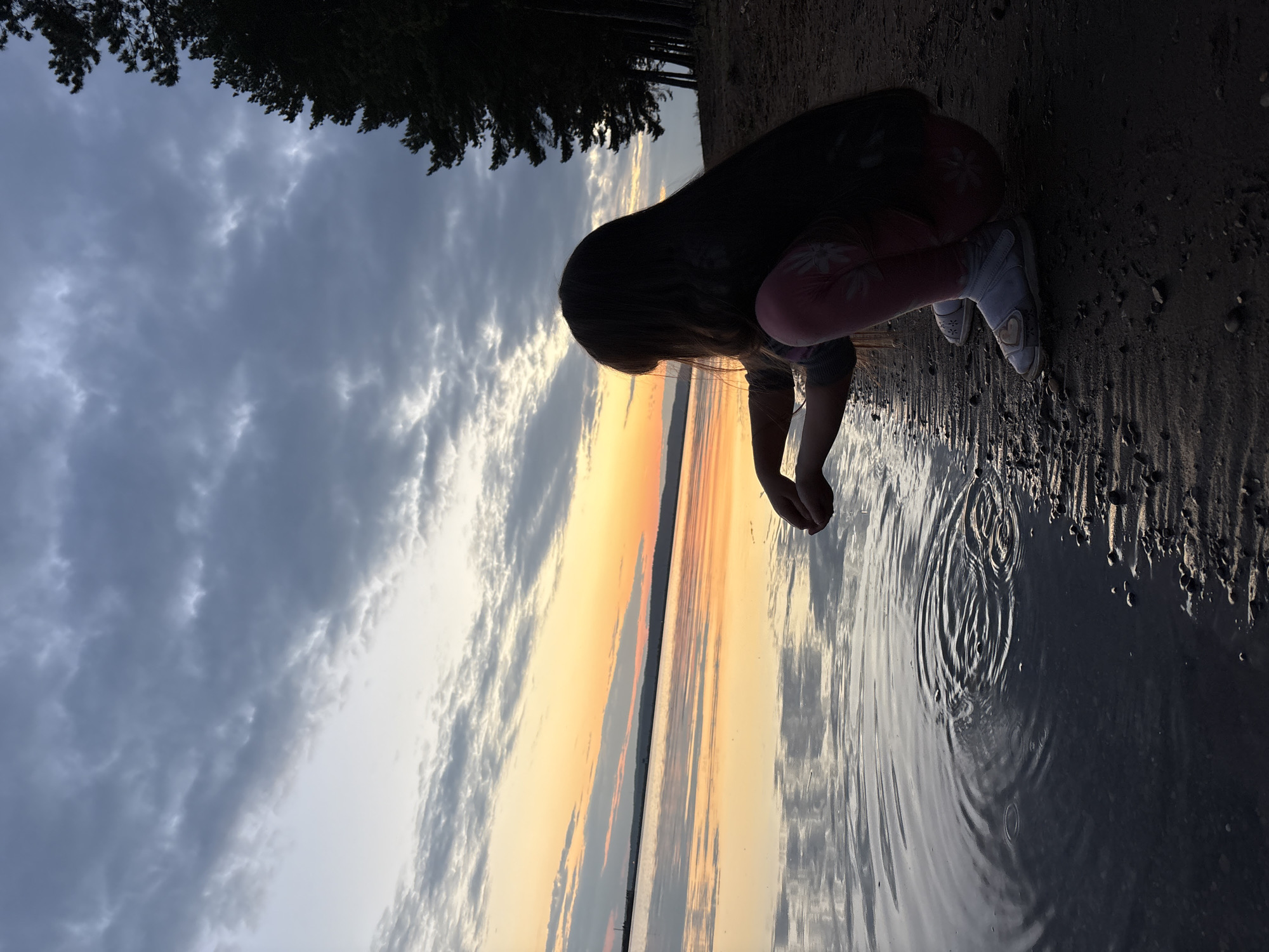 A young girl with long dark hair crouches on a sandy shore, her hands cupped as if to scoop water. She is wearing pink leggings with a floral pattern and white shoes. The girl faces a large body of water, likely a lake or ocean, where the sunset paints the sky with hues of orange and yellow. Dark trees line the right side of the image, silhouetted against the colorful sky. The water's surface is calm, with small ripples emanating from where the girl is interacting with it. The overall atmosphere is peaceful and serene, capturing a moment of quiet contemplation by the water at dusk.