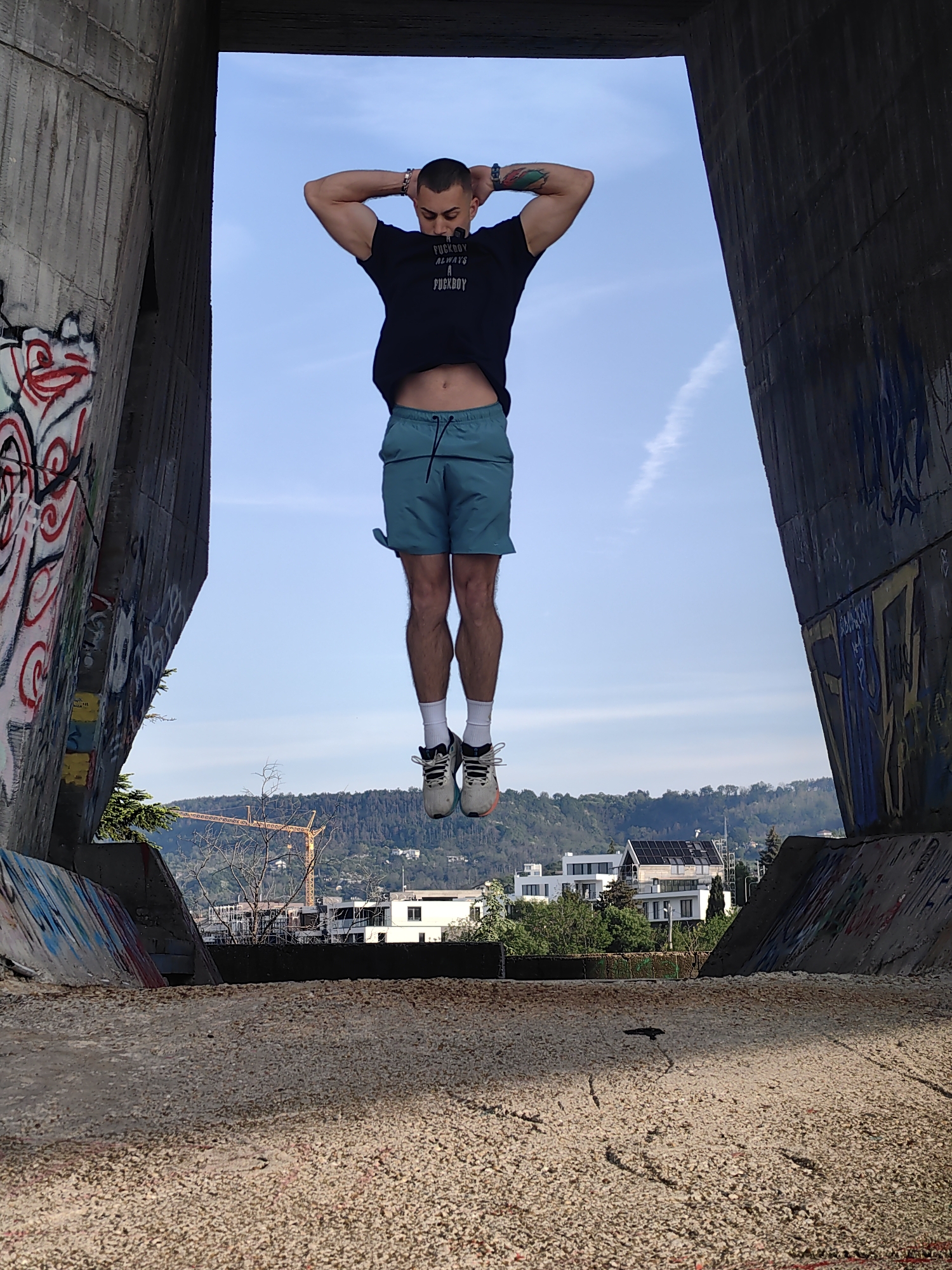 A man is jumping mid-air, seen from a low angle through a large concrete structure with graffiti on its sides. The man wears a black t-shirt, blue shorts, white socks, and grey running shoes. His hands are behind his head, and he appears to be posing during the jump. Beyond the concrete structure, there is a view of buildings, trees, and a hill with a yellow construction crane. The sky is light blue with a faint contrail. The ground beneath the structure is covered in small rocks.