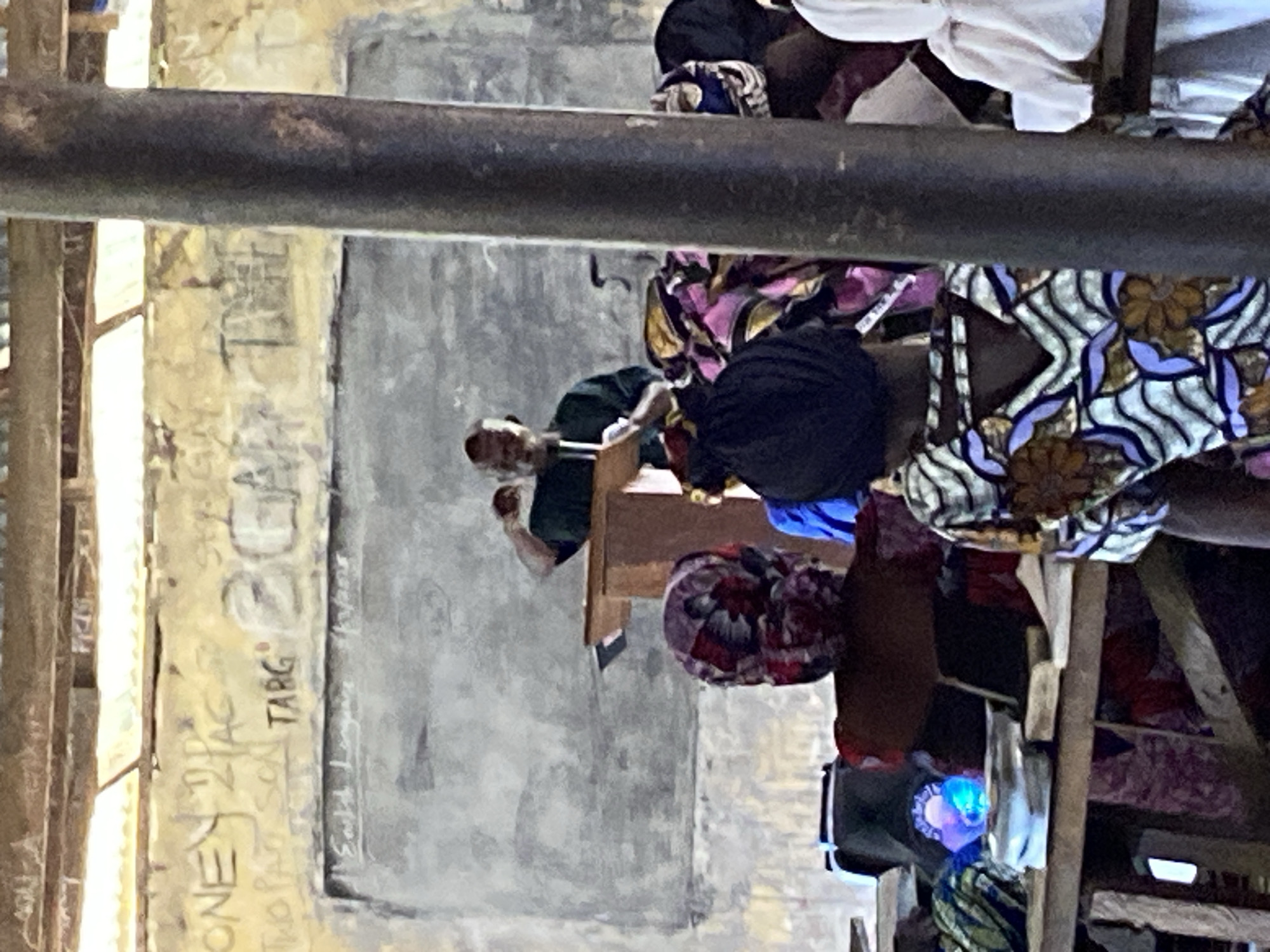 The image, taken indoors in Mararaba, Nigeria, shows a group of people seated. Some individuals are wearing colorful, patterned fabrics. A man is standing at a wooden desk. Graffiti and writing are visible on a nearby wall. The lighting appears to be a combination of natural light from a window and artificial light. The scene suggests a classroom or meeting setting.