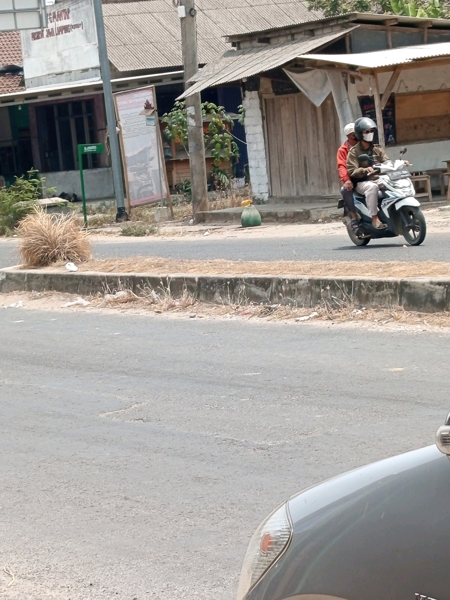 In Pulung Kencana, Indonesia, a motorcycle carrying two individuals passes by a building with a corrugated metal roof and a wooden facade. The driver, wearing a helmet and a face mask, is dressed in a brown jacket, while the passenger wears a red shirt. The motorcycle is white and has a license plate that reads "B 5774 ZAQ". A green roadside marker is visible to the left of the building. The road is paved, and a concrete median with dry vegetation separates it from the sidewalk. In the foreground, a portion of a gray car is visible, with its headlight and hood illuminated by sunlight. The scene appears to be a typical street view in a rural or suburban Indonesian setting.
