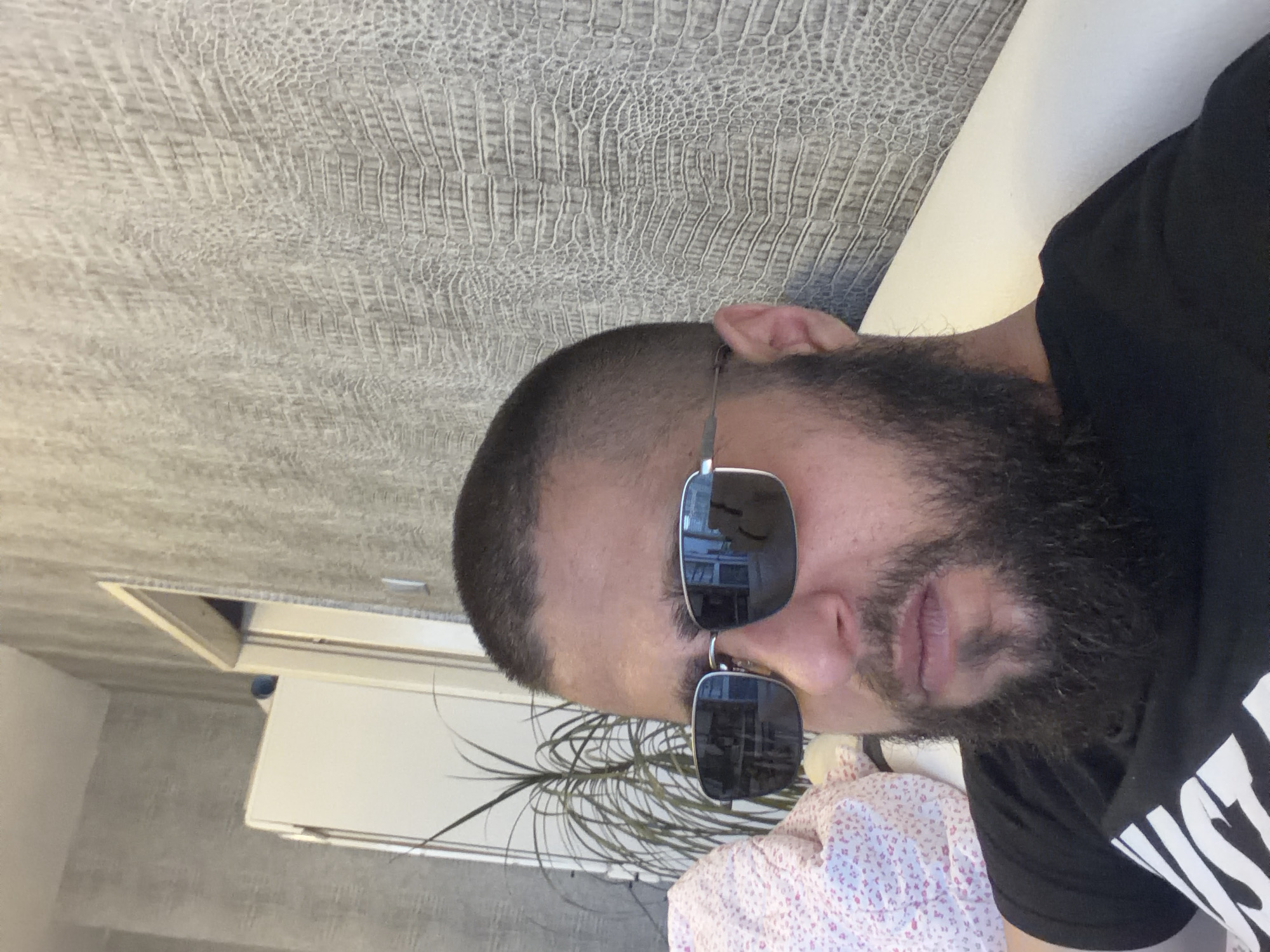 A man with short hair and a full, dark beard wears silver-framed sunglasses and a dark t-shirt. He is positioned at an angle against a wall with a textured, reptilian-like pattern. Behind him, a white cabinet is partially visible, along with the foliage of a houseplant. He appears to be lying on a white surface, possibly a bed or couch, partially covered by a pink and white floral patterned fabric. The location is an interior space of Unknown, Unknown.