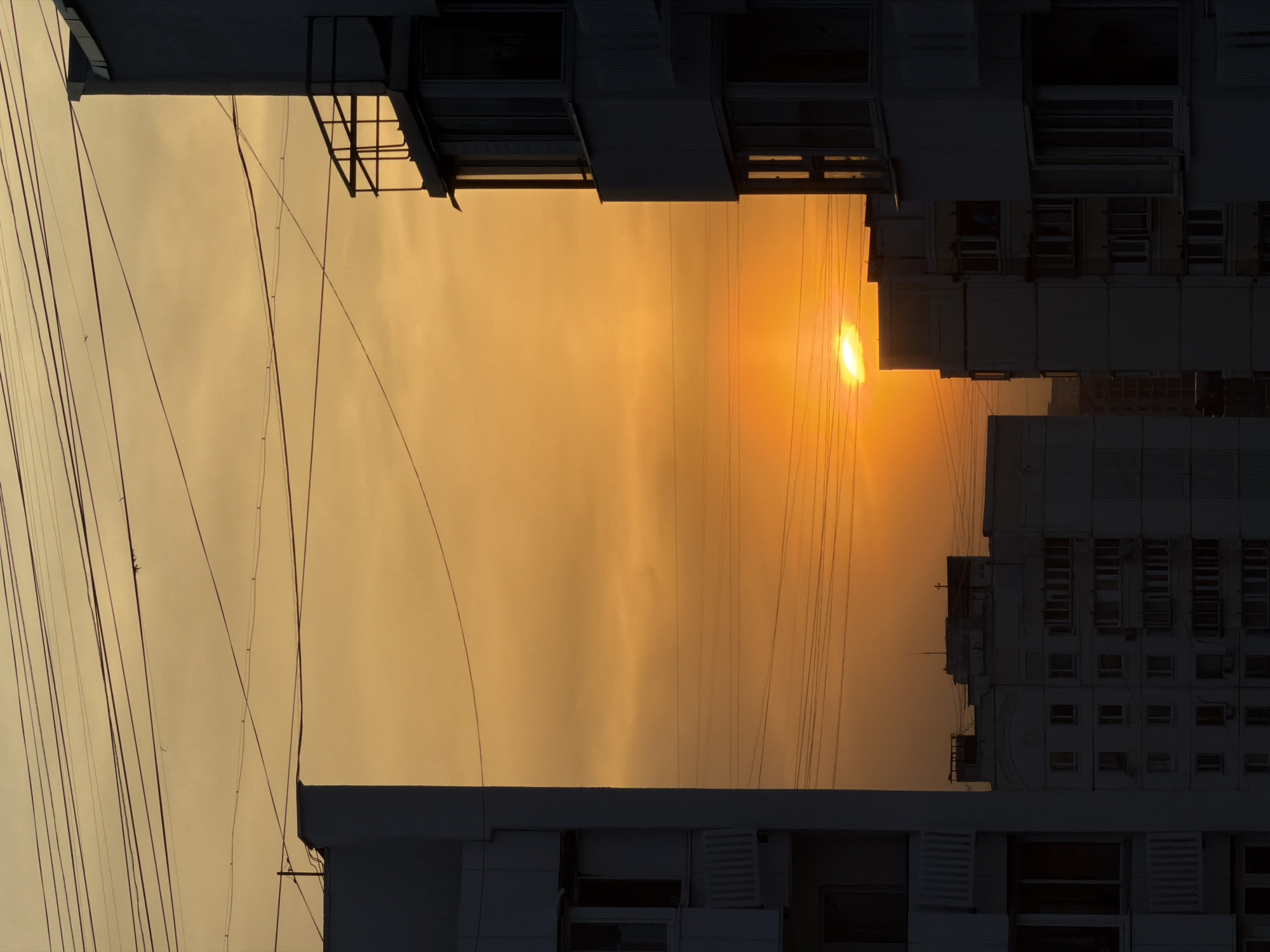 Captured in Khimki, Russia, the image presents a sunset viewed between buildings. A bright sun glows amidst an orange-hued sky, bisected by numerous power lines that stretch from the foreground to the horizon. Buildings stand prominently on both sides, their silhouettes defined by windows and architectural details. The sky appears overcast, contributing to the warm color palette.