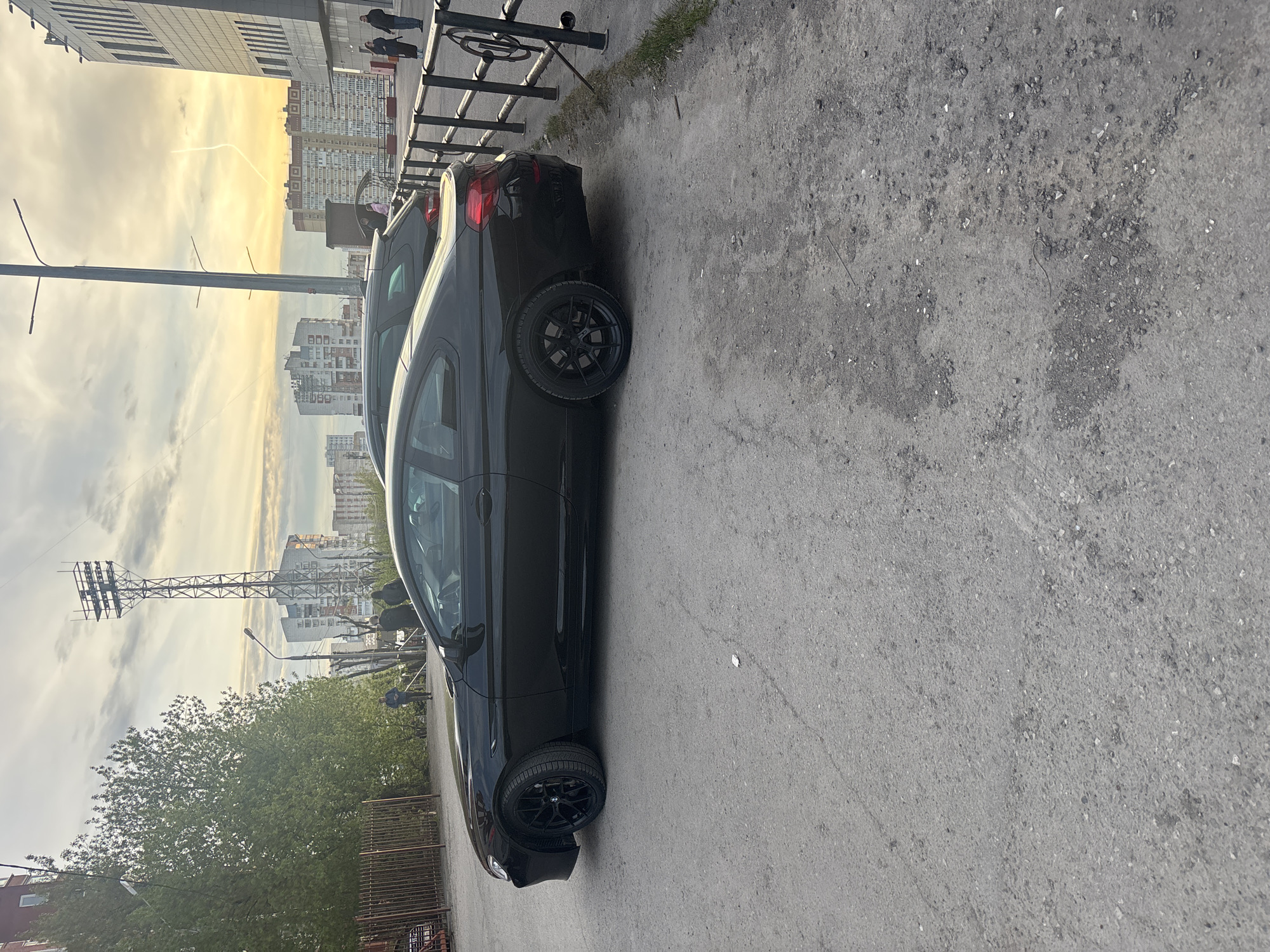 The image depicts a black BMW coupe parked on a gray asphalt road. A metal fence runs along the edge of the road, and pedestrians are visible walking along the sidewalk adjacent to the fence. In the background, buildings characteristic of a Russian city skyline are visible under a sky with clouds tinged orange by the setting sun. A power line tower is also visible. The setting is likely Perm, Russia, based on the architecture and infrastructure.