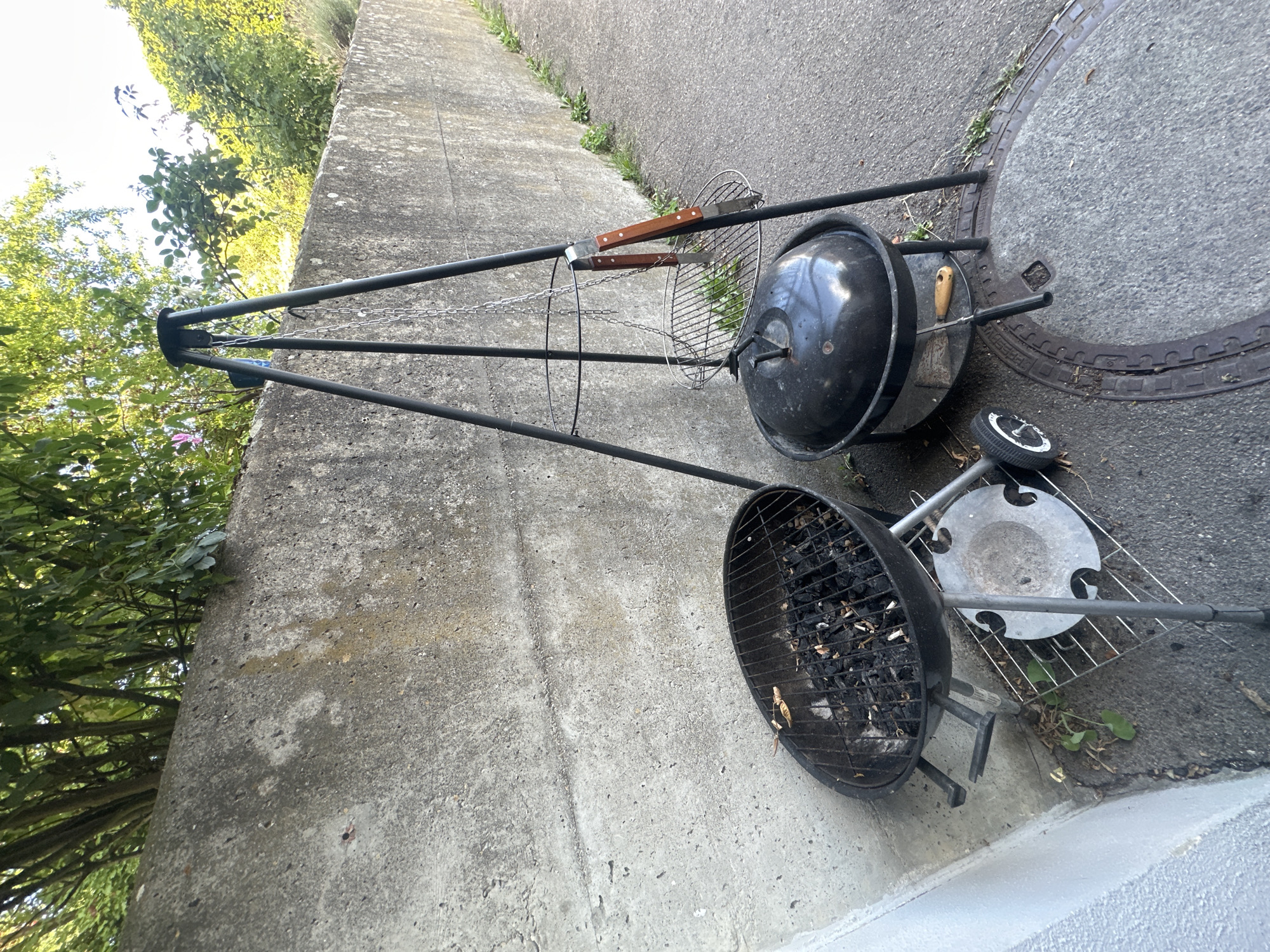 An outdoor scene on a paved surface, likely a sidewalk or alleyway, adjacent to a rough concrete wall. Two charcoal grills are present. The first, an oval-shaped grill with a black grate, sits on a stand with wheels and contains burnt charcoal. The second, a dome-shaped black grill with a lid, is positioned to the right of the first. Above the second grill is a black tripod stand with a chain hanging from its center. A pair of barbecue tongs with wooden handles rests on the tripod. Green foliage is visible at the top of the image, growing above the concrete wall. The scene is illuminated by natural daylight, suggesting an outdoor setting in a city like Günzburg, Germany.