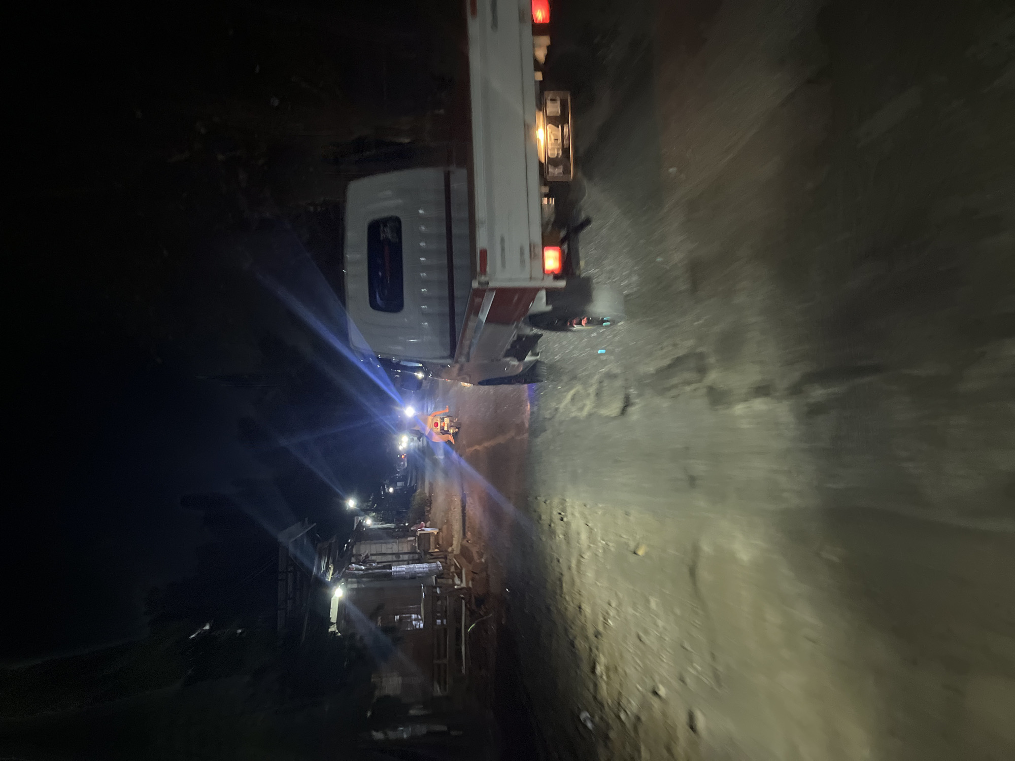 The image depicts a nighttime street scene in Pecangaan, Indonesia. A white utility truck with red accents is in motion on a dirt and gravel road. The truck's license plate is visible and reads "K 9672 UD". To the left of the truck, there are several dimly lit roadside structures that appear to be small shops or dwellings. Further down the road, a person is riding a scooter, illuminated by their own headlights. The road is uneven and appears to be unpaved. The scene is illuminated by the headlights of the vehicles and streetlights.