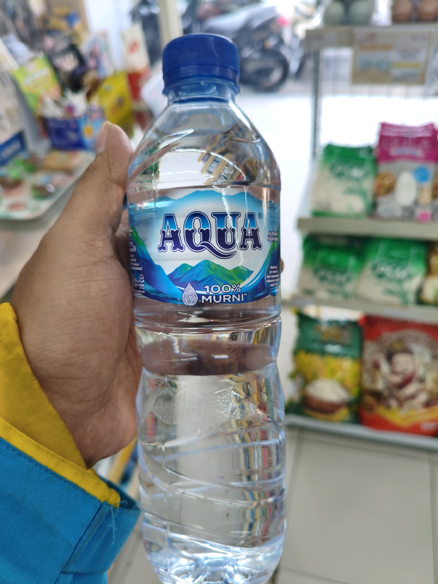 A close-up shot shows a hand holding a plastic bottle of Aqua brand mineral water. The bottle has a blue cap and a label featuring a mountain landscape and the brand name in large, blue lettering. Below the brand name, it reads "100% MURNI". The water inside the bottle is clear. In the background, shelves are visible, stocked with various packaged goods, suggesting the location is likely a convenience store or minimarket in the city of Sumberejo Kulon, Indonesia. The items on the shelves are blurred but appear to be snacks and other food products. The lighting suggests an indoor environment.