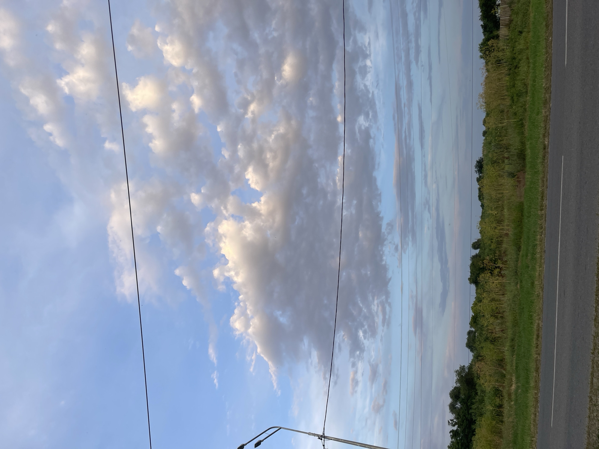 The image depicts a roadside scene in the city of Donskoye, Russia. The foreground features a paved road with a white line marking its center. Beyond the road lies a strip of grass, followed by a dense line of green vegetation and trees that form the horizon. Above the vegetation, several parallel power lines stretch across the frame. The sky is a blend of blue and white, with scattered cumulus clouds illuminated by the sun, suggesting it is either late afternoon or early morning. A street lamp with two fixtures is visible on the left side of the image, along with its supporting pole and attached wires. The overall impression is one of a rural or semi-urban environment.