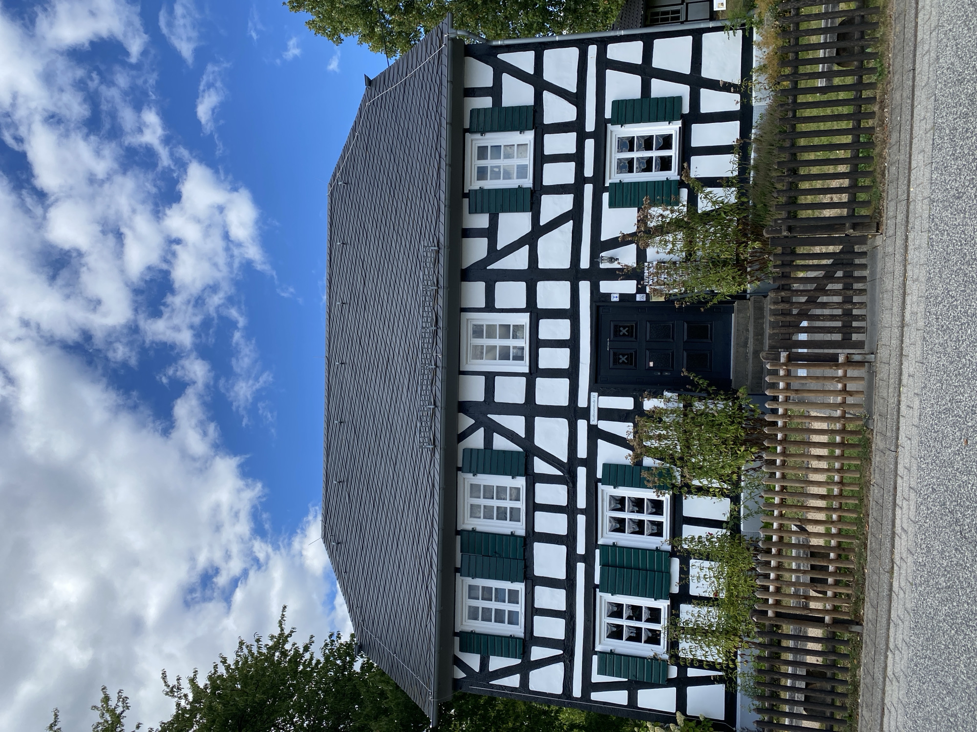 The image shows a two-story half-timbered house with a dark gray shingle roof, located in Flammersfeld, Germany. The facade is a traditional black and white timber frame construction with white infill. There are multiple windows with green shutters on both floors. A dark-colored front door is centrally located on the ground floor, accessed by a short set of stairs. In front of the house, there is a wooden picket fence and some vegetation. The sky above is blue with scattered white clouds. The scene is a street view with asphalt pavement in the foreground. No people are visible in the image.