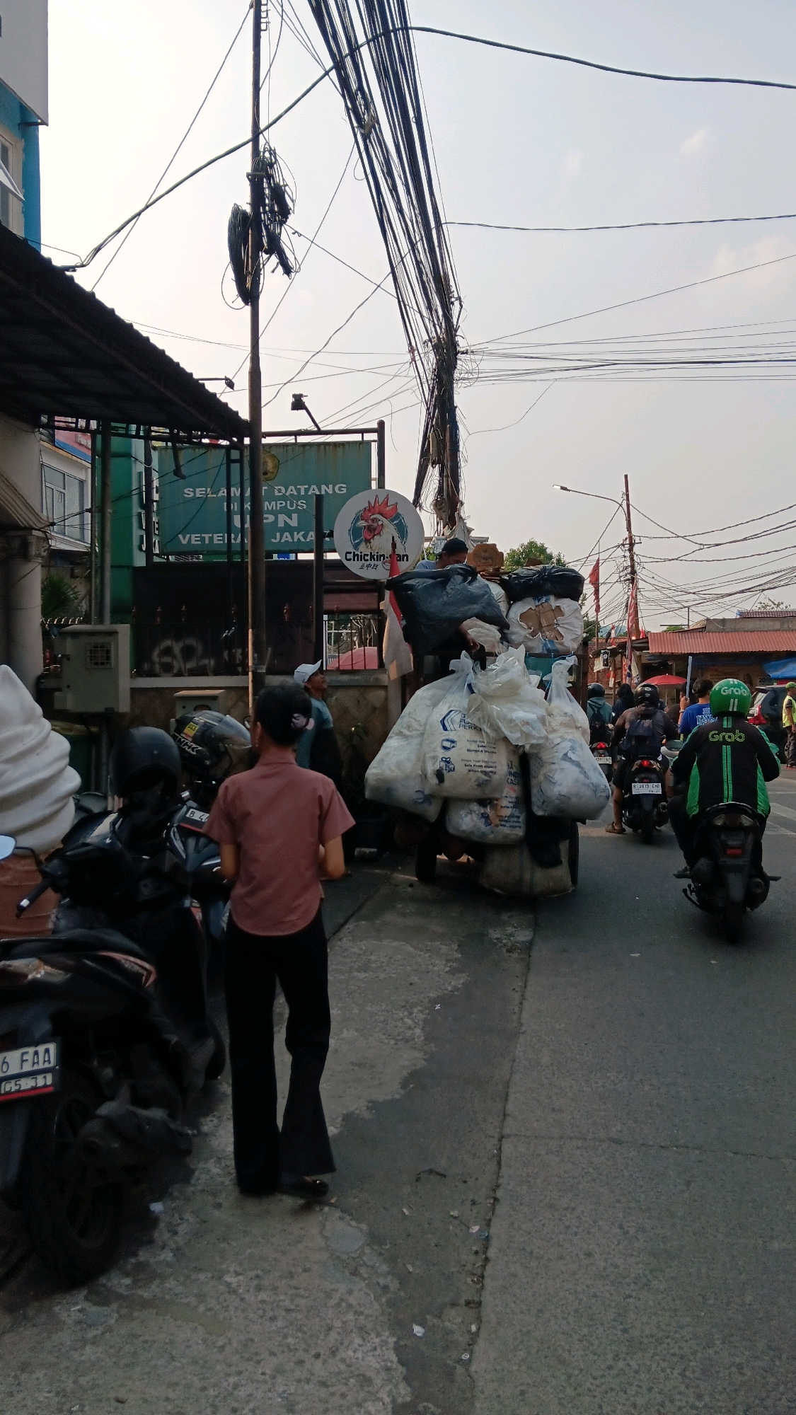 This image depicts a street scene in Depok, Indonesia, likely in an urban or semi-urban area with significant overhead wiring. In the foreground, a person wearing a pinkish-brown short-sleeved shirt and dark pants stands facing away from the camera, with their back to a pile of large plastic bags filled with what appears to be recyclables or waste. To the left, several parked motorcycles are visible, one with a license plate that reads "B 46 FAA".
Behind the standing person and the pile of bags is a cart, possibly a handcart or a makeshift trailer, overloaded with more bags and other items. A sign with Indonesian text, including "SELAMAT DATANG" (Welcome) and "UPN VETERAN JAKARTA", is visible on a building to the left, suggesting the location is near this university. There's also a logo with a chicken on it, possibly for a food establishment.
Further down the street, several individuals on motorcycles are present. One motorcycle is ridden by a person wearing a bright green jacket with "GRAB" prominently displayed, indicating a ride-sharing service. Other motorcycles and people are seen in the background, with more buildings and shops lining the street. The sky is overcast, suggesting daytime with possibly hazy conditions. The overall impression is one of a busy street with local commerce and daily activities taking place.