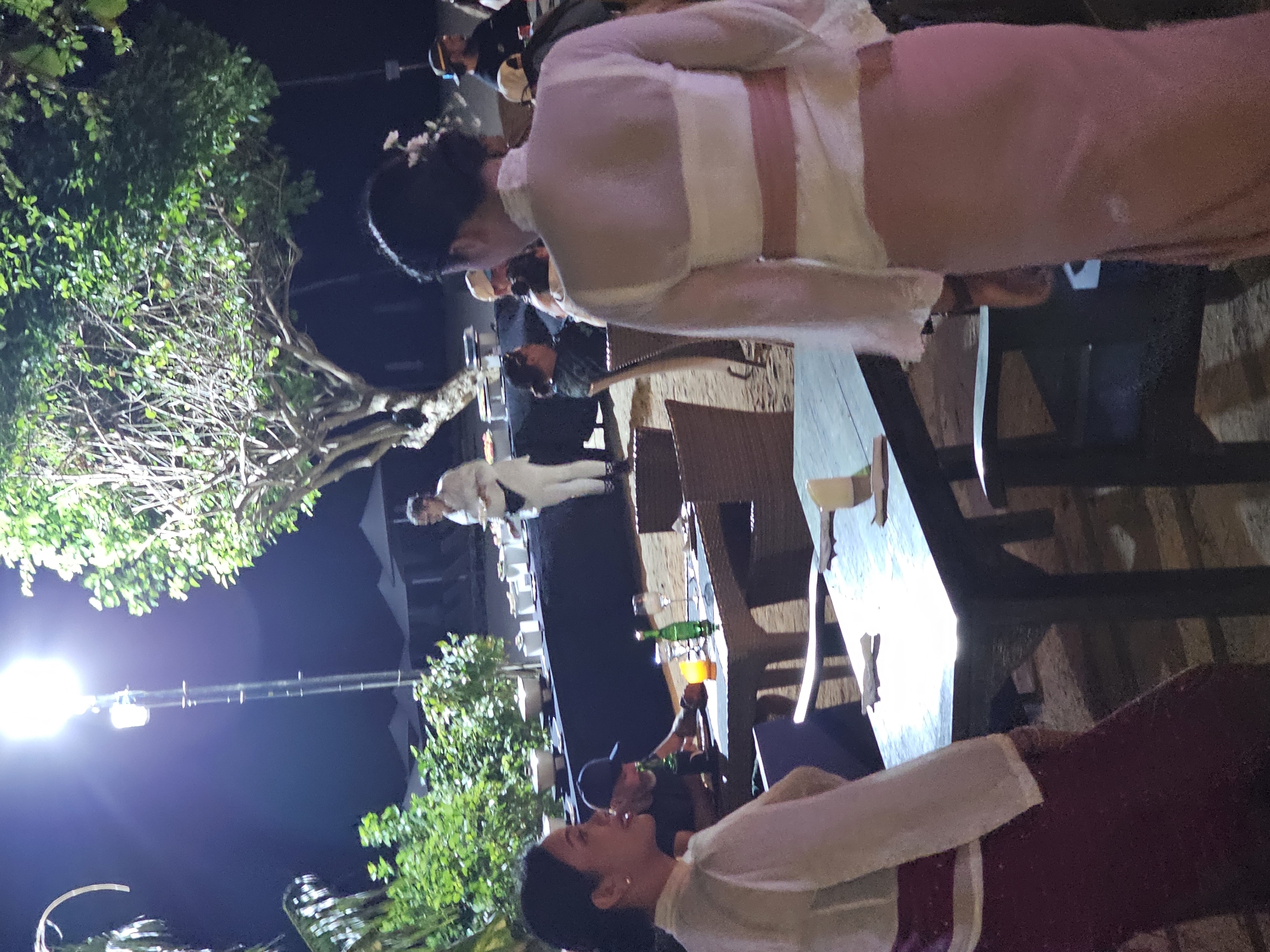In Denpasar, Indonesia, at night, a group of people are gathered at an outdoor dining area, possibly a restaurant or resort. The scene is illuminated by a bright overhead light and ambient lighting, with lush greenery and trees surrounding the area. In the foreground, two women are visible. The woman on the left is wearing a traditional outfit with a white sheer top over a maroon skirt, and she is looking to her left. The woman on the right is facing away from the camera, wearing a similar white sheer top with a pink sash and a long pink skirt.
Several tables are set up, some with place settings and a bottle of wine, suggesting dining is taking place. In the background, a buffet line is set up with various food containers, and a server in a white uniform is attending to it. Other guests are seated at tables or standing, engaged in conversation. The ground appears to be sandy, indicative of a beachside location. The overall atmosphere is that of an evening event or gathering in a tropical setting.