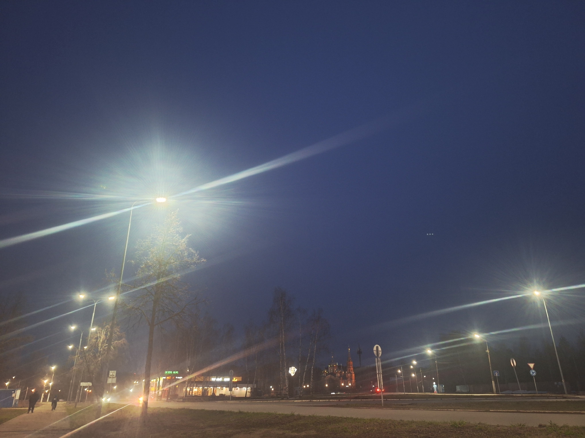 The image depicts an urban landscape at night in Chaikovsky, Russia. Several street lights illuminate the scene, creating lens flares and visible light rays in the dark sky. The sky is a deep blue with a few faint stars or possibly distant lights visible.
On the left side of the image, a sidewalk with two pedestrians walking away from the camera is seen. A row of streetlights lines the sidewalk. On the right, across a road, is a building with illuminated windows, possibly a restaurant. In the distance, an Orthodox church with multiple domes and towers is visible. Various road signs and traffic lights are scattered along the road. The foreground shows a patch of grassy area.