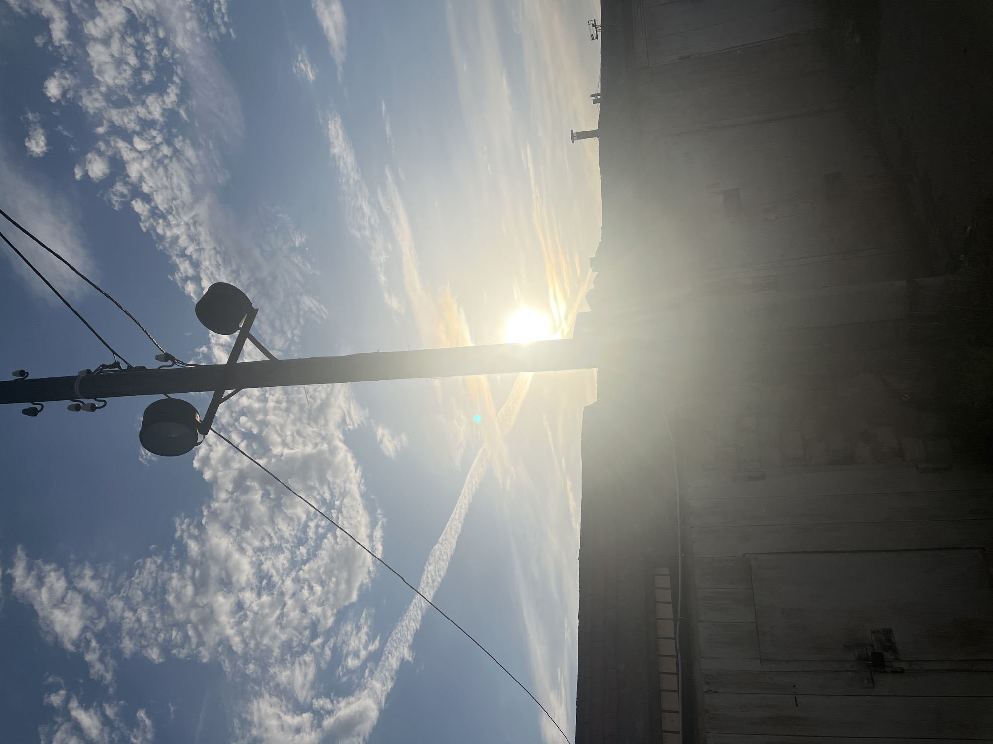 The image depicts a sunlit urban scene in Chaikovsky, Russia. A utility pole with two streetlights is visible in the left foreground, extending upward into a blue sky with scattered clouds and contrails. A building abuts the right side of the frame, largely obscured by glare. The sun is positioned centrally, causing significant lens flare. The overall impression is bright and overexposed.