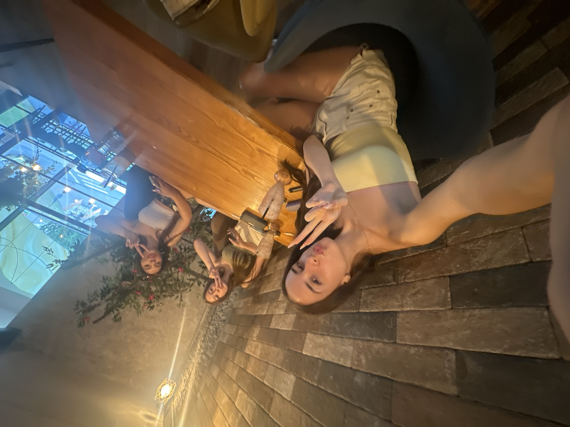 In Shumen, Bulgaria, at a restaurant with exposed brick walls and a wooden table, three young women are seated. The woman closest to the camera, in the foreground, is taking a selfie, holding up two fingers in a peace sign and puckering her lips. She is wearing a strapless yellow top and light-colored shorts. To her left, another woman in a green top also holds up two fingers in a peace sign. Further to the right, a third woman in a white top is partially visible, also making a peace sign. A glass with a straw, a bottle of water, and a smartphone are on the table. The restaurant has a glass ceiling, with greenery visible outside.