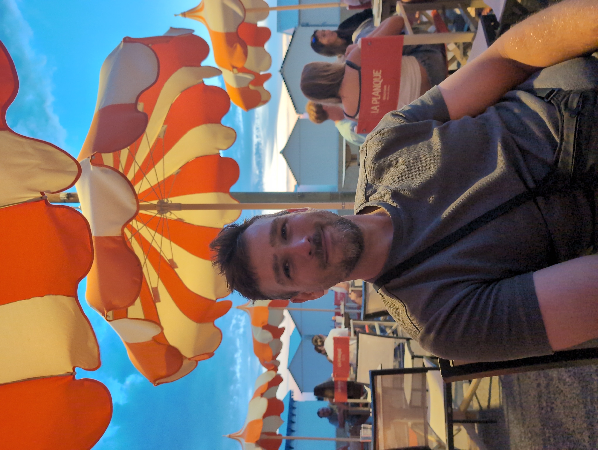 A man with a beard is smiling at the camera, seated outdoors under orange and white striped beach umbrellas in Mers-les-Bains, France. The sky above is blue with scattered clouds. In the background, several beach huts are visible, as well as other people seated at tables. One person in the background is seated on a red chair with the words "LA PLANQUE" printed on it. The man is wearing a grey t-shirt and has a dark strap across his chest. The scene appears to be a beachside cafe or restaurant.