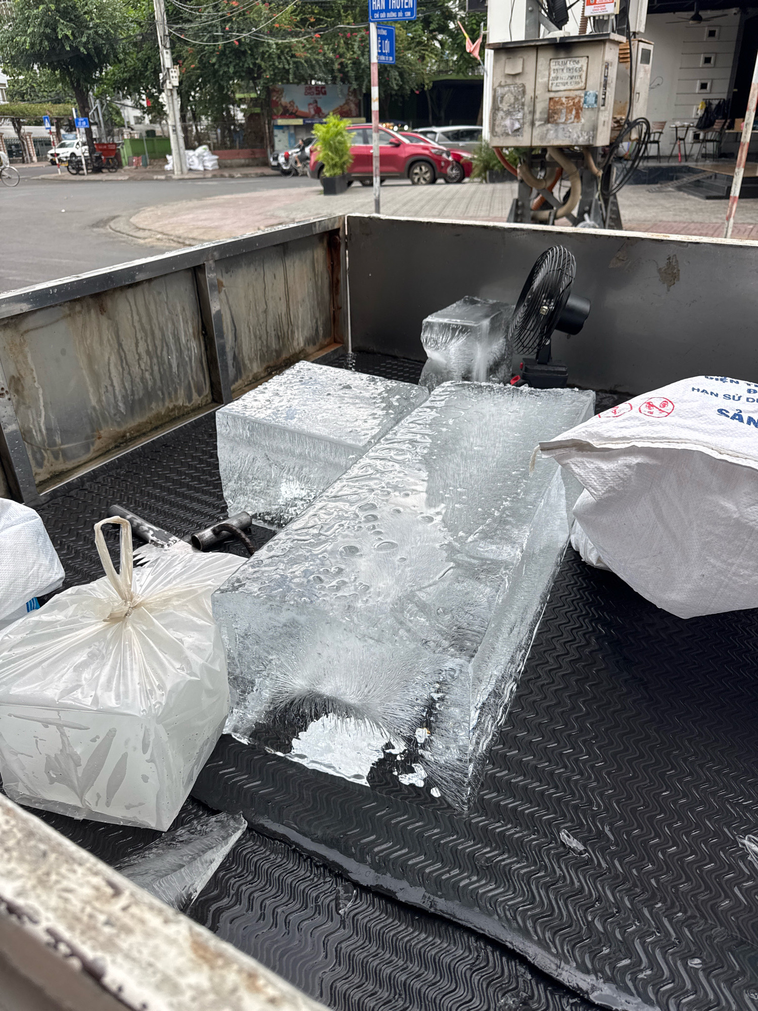 The image shows the back of a truck bed filled with large blocks of ice. A black electric fan is positioned near the ice blocks, possibly to keep them cool. To the left of the ice is a white plastic bag, and to the right is a white woven bag with Vietnamese text printed on it. The truck bed has a black, textured surface. In the background, the scene appears to be a street in Nha Trang, Vietnam, with buildings, trees, a traffic light, and vehicles visible. The presence of the ice and fan suggests it's a delivery or preparation for an event where ice is needed.