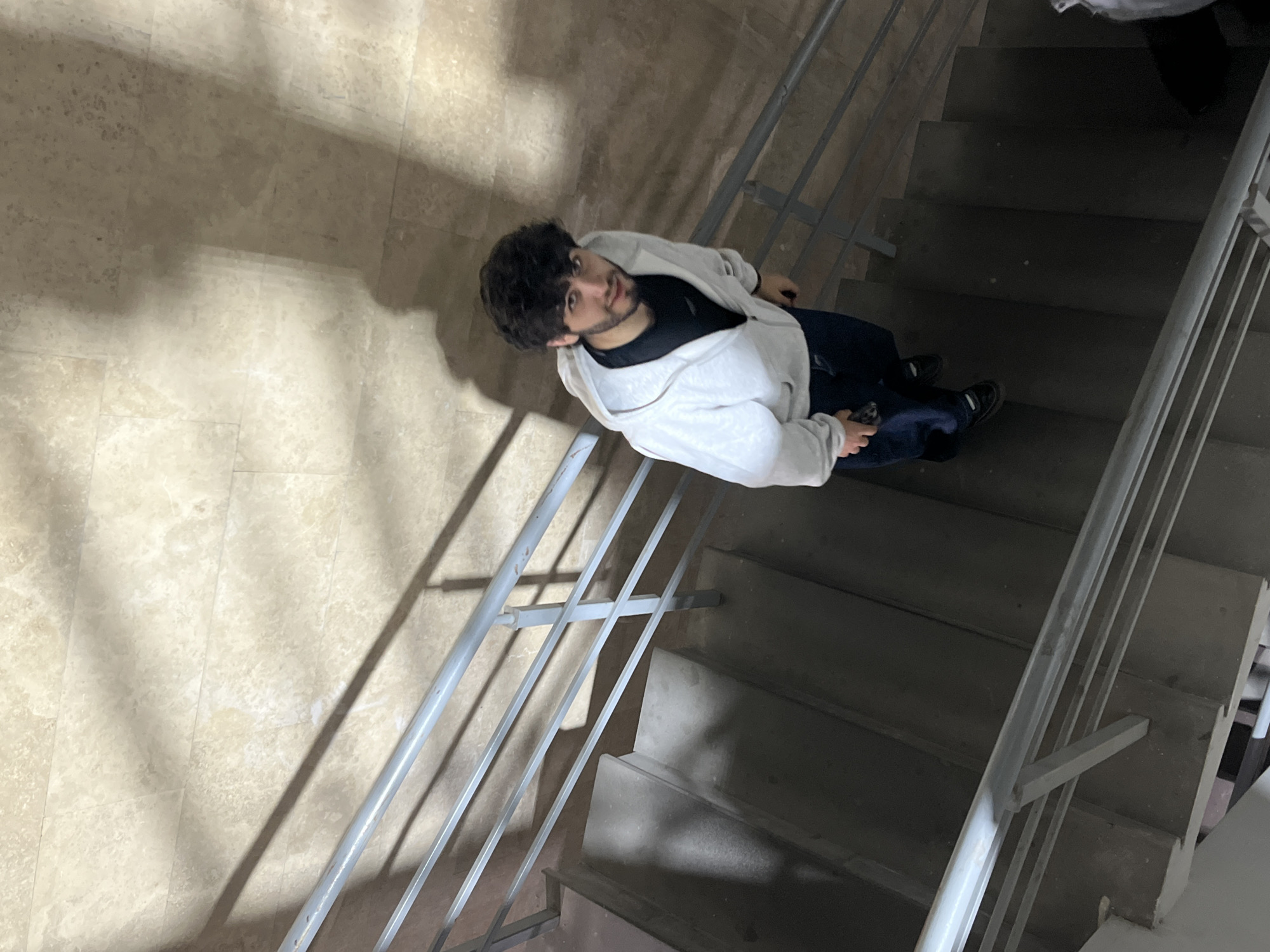 The image depicts a man with dark curly hair and a beard, wearing a black shirt, dark jeans, a light-colored hoodie, and black shoes, standing on a staircase in a building. The staircase has a gray metal railing. The walls are covered in light-colored stone tiles. No information is available to determine the exact location in Yerevan, Armenia.