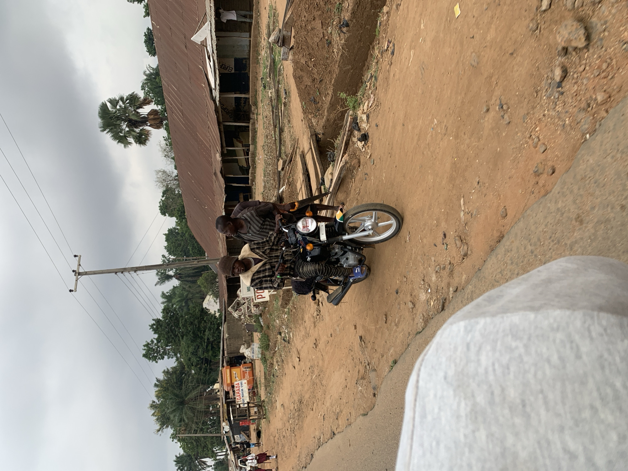 Two men are riding on a motorcycle on a dirt road in what appears to be Maigizo, Nigeria. The man in front wears a striped shirt, while the other man wears a plaid shirt and a hat. Buildings line the road, and there are trees in the background. A pole with electrical wires is visible. The lighting suggests it is daytime. There is a business sign that says GAHADA BEST PROVISION STORE.