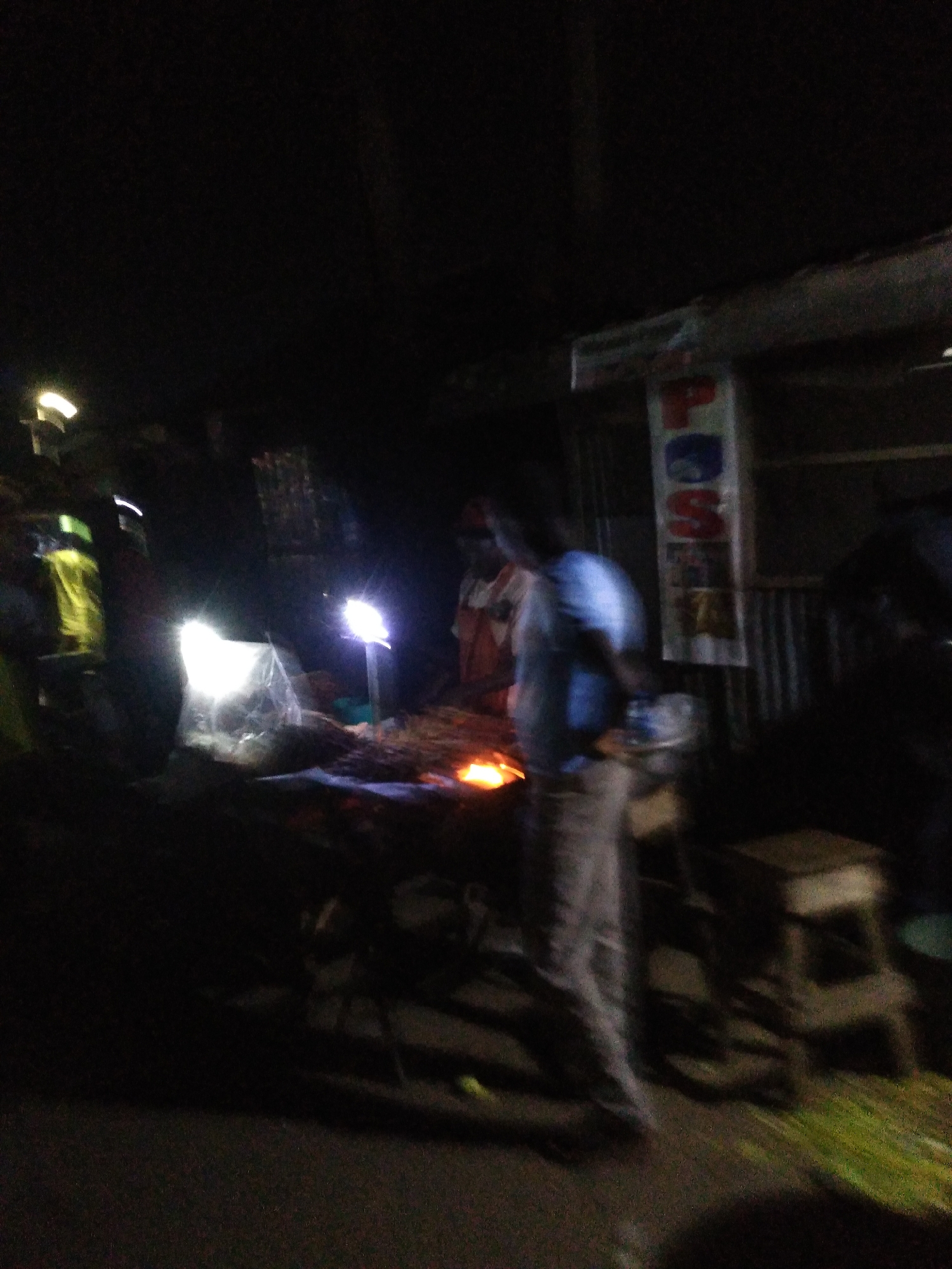 This is a nighttime, low-light, slightly blurred photograph of an outdoor market stall in Gura Top, Nigeria. A vendor, wearing a blue shirt and light-colored pants, stands behind a stall displaying food, possibly grilled meat. The stall is illuminated by a bright light, creating strong highlights and deep shadows. In the background, there is a sign with red and white lettering, partially obscured and unreadable. Another individual, wearing a brightly colored yellow garment, is visible to the left. The ground appears to be dirt or gravel, and there are various items on display or stored in the vicinity, including what looks like bundles of grass or foliage in the foreground. The overall atmosphere is busy and functional, typical of a nighttime market.