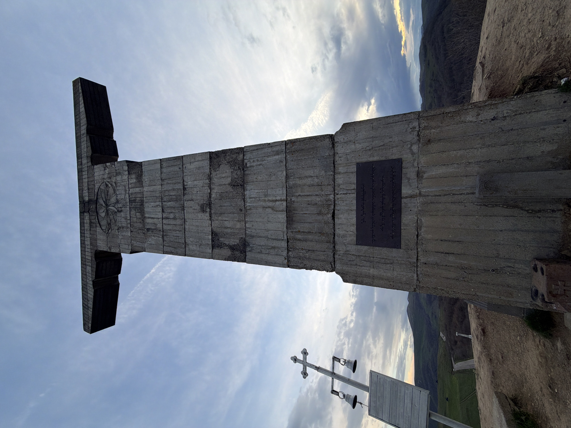The image depicts the Monument of Georgian Alphabet in Zemo Nichbisi, Georgia. The monument is a large concrete structure with a T-shape at the top and a relief of an ancient Georgian symbol on the front. A plaque with text is affixed to the side of the monument. The surrounding landscape includes mountainous terrain and a cloudy sky. In the foreground, there is a cross-shaped bell tower and a wooden sign. The monument stands on a cliff edge with vegetation nearby.