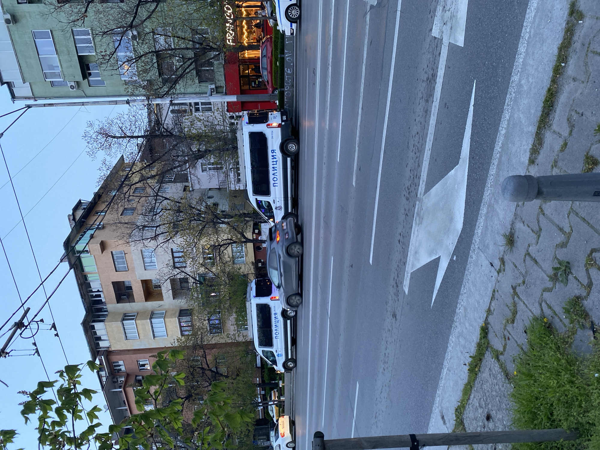 The image depicts a street scene in Sofia, Bulgaria, featuring two police vans parked in the middle of a multi-lane road. A grey sedan is also visible between the two vans. On the side of the road, there's a sidewalk with a metal bollard. Arrow markings on the asphalt indicate the flow of traffic. Buildings of varying heights and architectural styles line the street, including one with a sign that reads "FRANCO". Overhead cables run along the street. The sky is overcast. The overall scene suggests an urban environment.