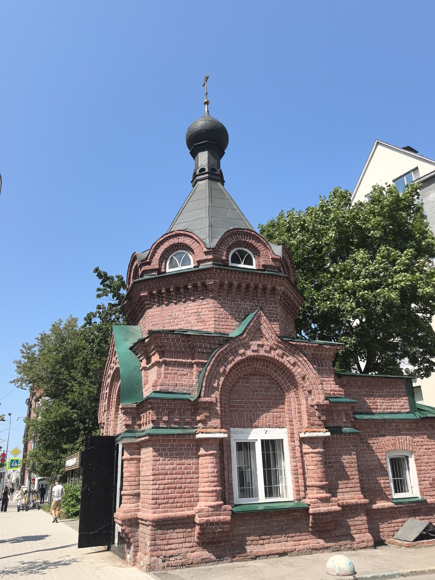 The image shows a small, red brick Orthodox chapel with a dark, onion dome topped with a cross. The chapel has arched windows with white frames and green metal awnings. A large, black metal door is partially open on the left side of the chapel. The building is situated on a paved sidewalk in front of a street with a few people walking and riding electric scooters. In the background, there are trees and a modern, white building. The sky is clear and blue. This chapel is likely located in Nizhny Novgorod, Russia, given the architectural style and the presence of Cyrillic text on a street sign visible on the left.
