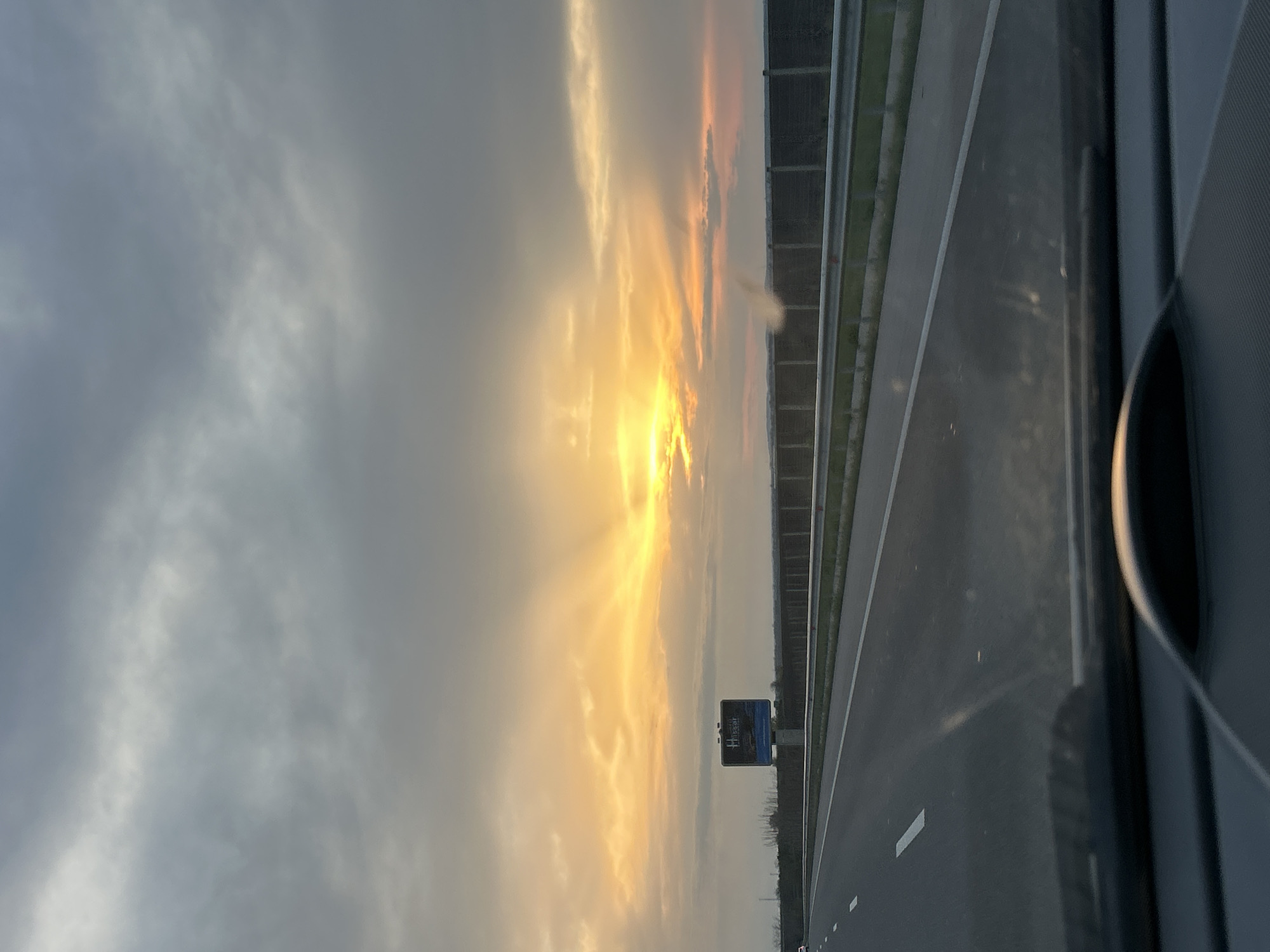 The image shows a view from inside a vehicle traveling on a highway. The sky is predominantly cloudy with the sun setting or rising, casting orange and yellow light through the clouds. A highway sign is visible next to the road. The road surface is grey asphalt with white lane markings and guardrails are seen beside the road. The image appears to be taken from the passenger seat. There is no other information that indicates the location is in the city of Cherna Gora, Bulgaria.