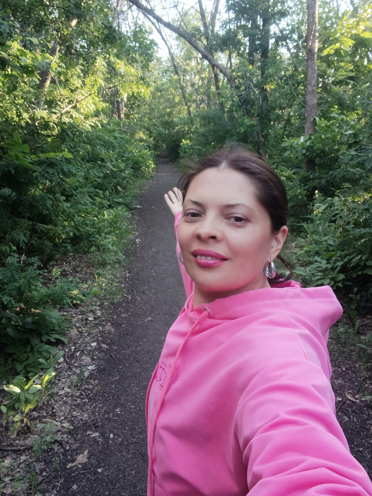 A woman with dark hair and dark eyes is captured in a selfie on a wooded path. She is wearing a bright pink hooded sweatshirt and large silver hoop earrings. Her left arm is extended, and her hand is raised, possibly waving. The path ahead of her is narrow and appears to be unpaved, flanked by lush green trees and undergrowth. The lighting suggests it is daytime, with sunlight filtering through the leaves. The environment is natural and appears to be a park or forest area. There is no indication of the city of Saratov, Russia, within the image itself.