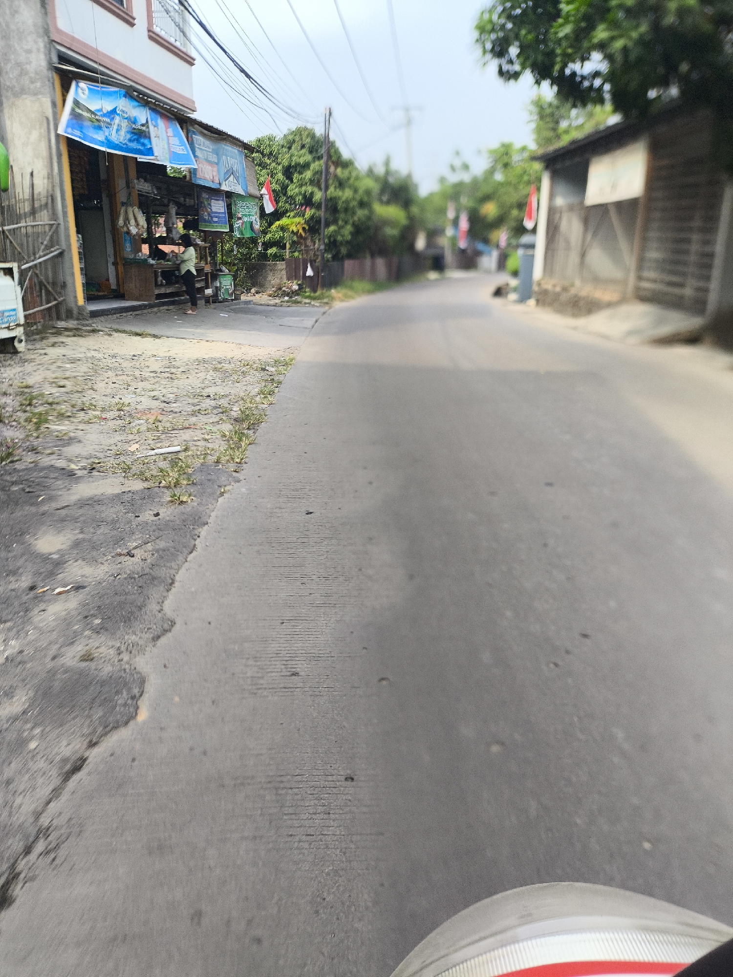 The image depicts a street scene in Cilegon, Indonesia. On the left side of the road is a small shop with banners displayed outside. A person wearing a light green shirt and dark pants is standing in front of the shop. The road itself appears to be paved and slightly worn. The background shows trees and other buildings, suggesting a semi-urban or suburban environment. The sky is overcast. The perspective suggests the photo was taken from a moving vehicle, likely a motorcycle, given the partial view of the front of the vehicle in the lower right corner.