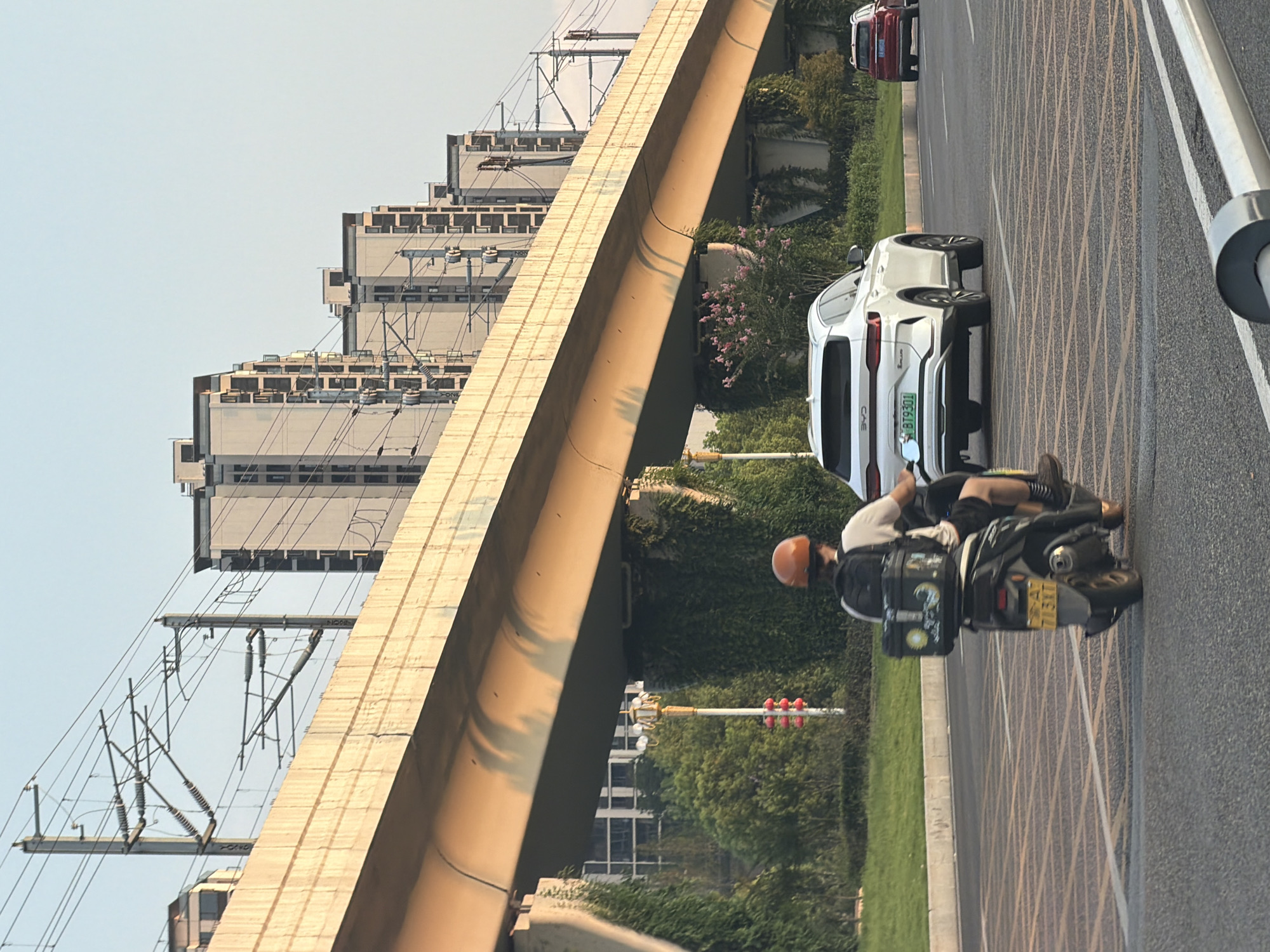 In Jiangjin, China, an overhead railway viaduct casts shadows on a road where a person on a scooter, wearing an orange helmet and a backpack with a black storage box, is moving away from the camera. The scooter has a license plate that reads "713RT". To the right, a white SUV with a license plate "BT9301" is in motion, followed by a red vehicle. Behind the road, buildings with multiple stories are visible, along with trees and vegetation. The sky is clear and bright, suggesting daylight.