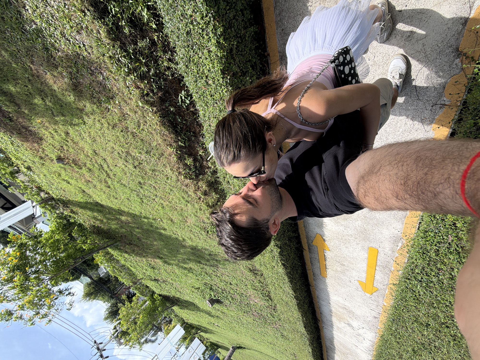 A couple is kissing on a sidewalk in Choeng Thale, Thailand. The woman is wearing a white dress, pink top, sunglasses, and white sneakers. The man is wearing a black shirt and khaki pants. The sidewalk is concrete with yellow directional arrows. There is a green hedge on the left side of the sidewalk and grass beyond. The sky is visible in the upper left corner.