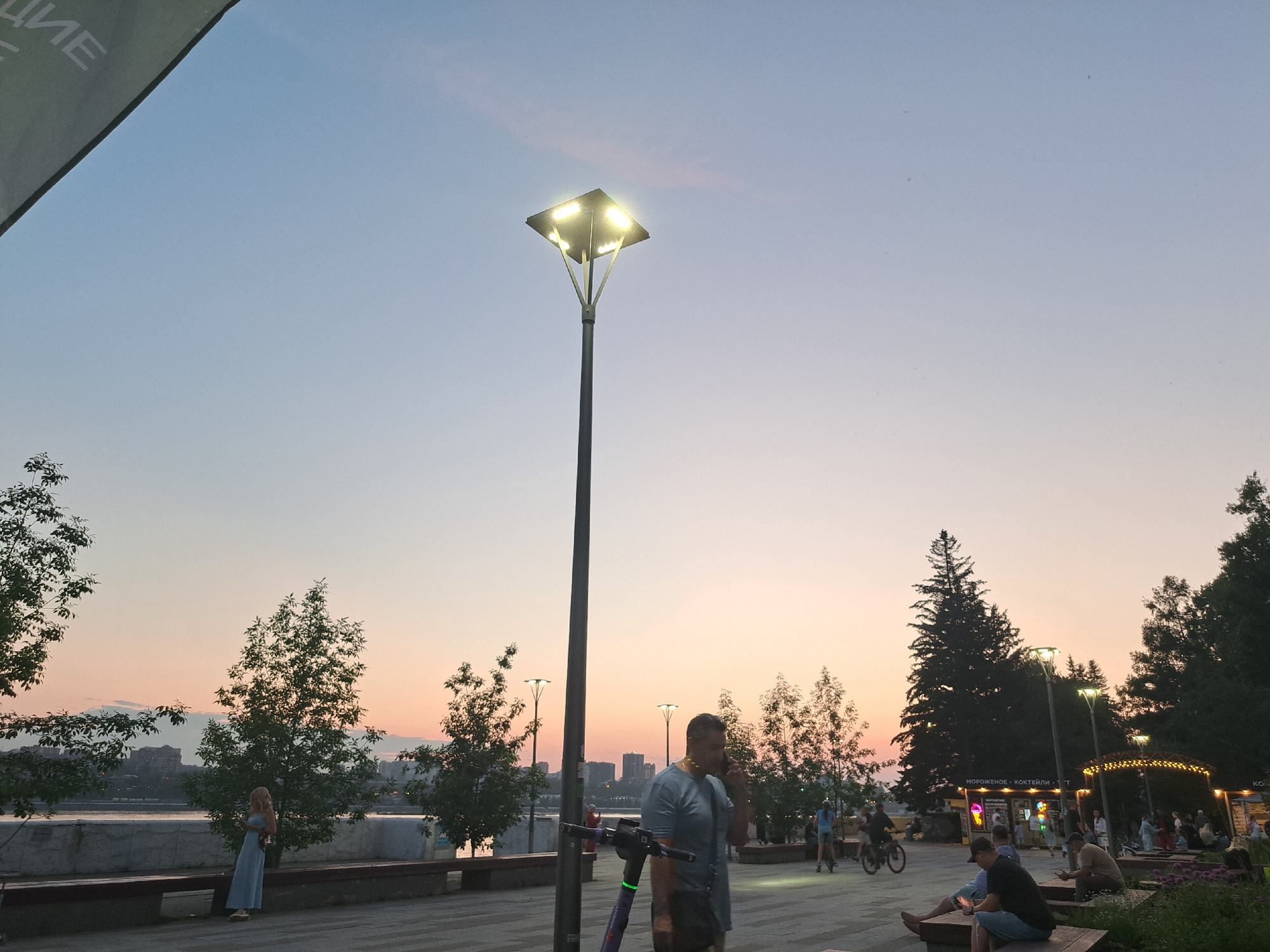 This is a horizontal, eye-level, medium shot of a park area in Irkutsk, Russia, during twilight. A tall, modern lamppost with four illuminated lights dominates the center of the frame. The sky is a gradient of light blue and pale pink, indicating sunset or sunrise. In the foreground, a paved pathway is visible, with several people scattered throughout.
To the left, a woman in a light blue dress stands near a tree, facing away from the camera. Further left, a large dark green structure, possibly an awning, is partially visible at the top left corner of the image.
In the mid-ground, a man in a grey t-shirt is walking and talking on his phone, with a purple electric scooter next to him. Behind him, a group of people are gathered, some sitting on benches and others standing. There are trees lining the walkway, and in the distance, buildings and a body of water are visible.
To the right, several more people are seated on benches and walking. A cyclist is visible on the right side of the image. In the background, more trees and a dark silhouette of buildings suggest a city skyline. Some businesses with illuminated signs are present in the distance, suggesting a commercial area within or adjacent to the park. The overall atmosphere is calm and relaxed, with the ambient light from the lampposts and signs casting a soft glow.