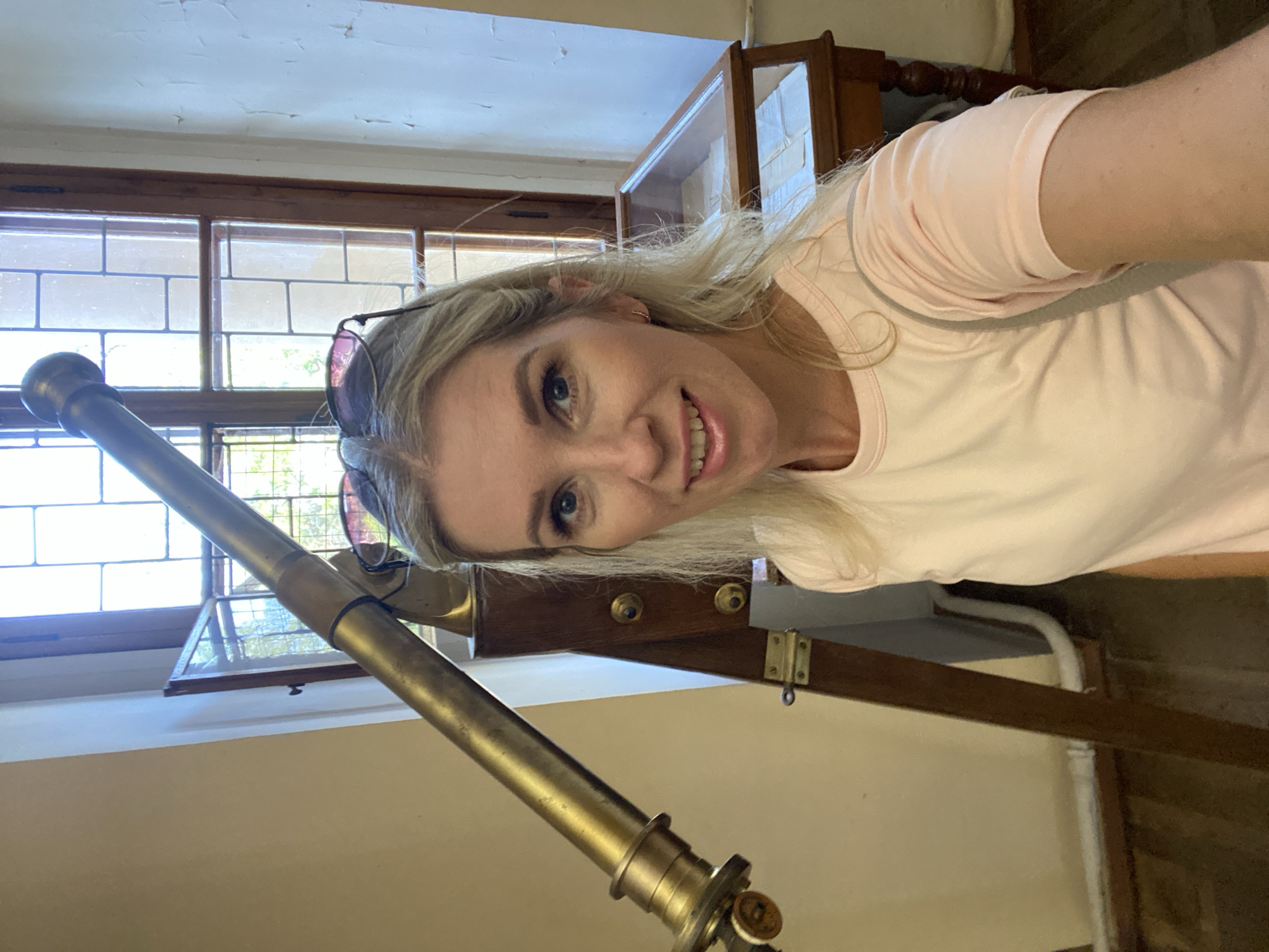 This is a portrait-style selfie taken by a woman in what appears to be an indoor setting with natural light. The woman has blonde hair and is smiling at the camera, wearing a pale pink t-shirt. She is also wearing sunglasses, which are pushed up onto her head. To the left of the woman is a brass telescope, and behind her, a large window with a grid pattern, suggesting it might be a stained-glass window. There are also wooden cabinets or furniture visible in the background, one of which has glass doors. The lighting suggests it is daytime. The overall mood of the image is casual and happy. No text is visible in the image.