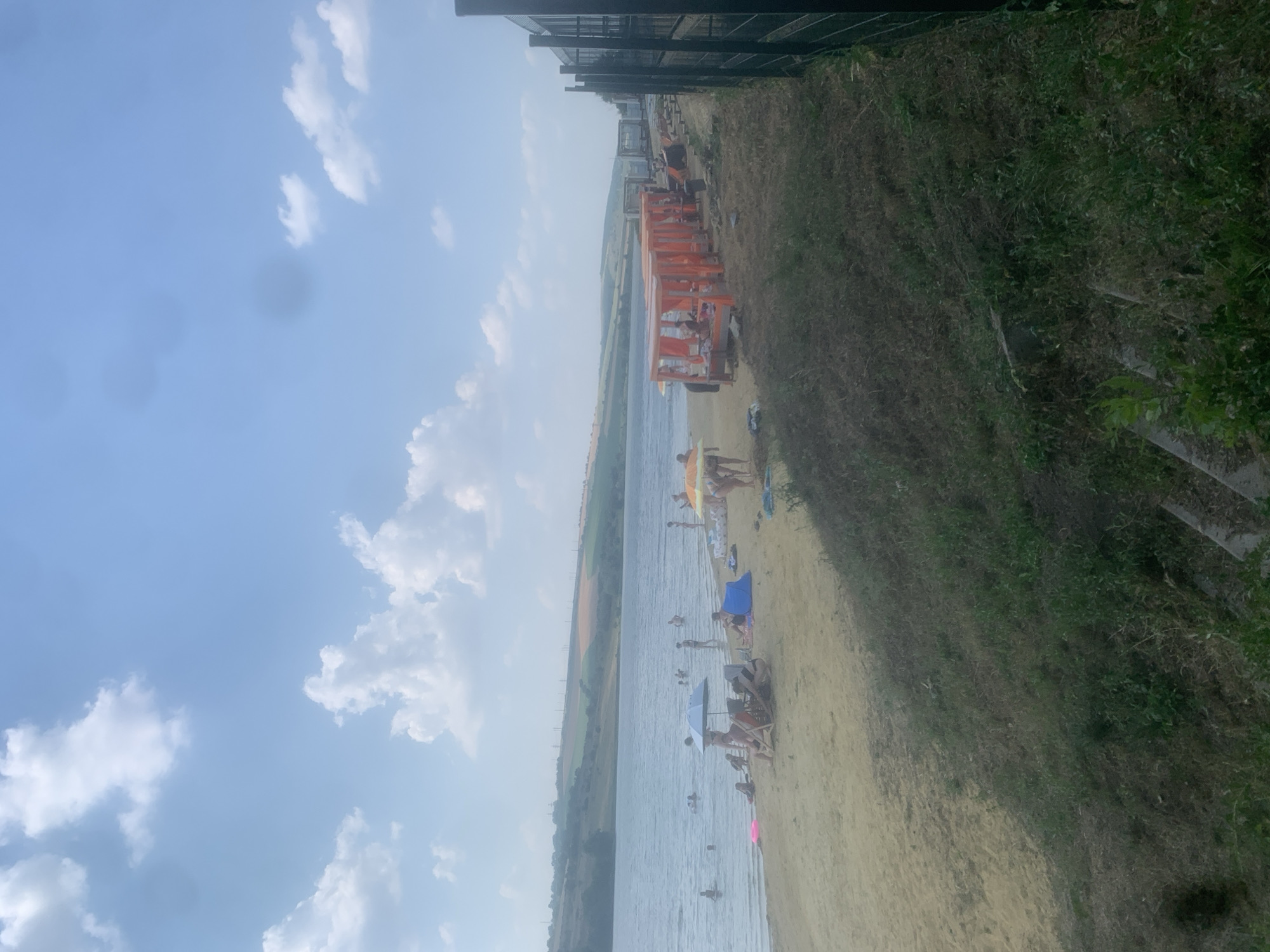 This image captures a sunny day at a beach with a calm body of water. Several people are visible enjoying the beach and water, with some sunbathing on lounge chairs and others swimming. An orange structure, possibly a canopy or seating area, is prominent on the beach, with people gathered underneath. Several umbrellas provide shade. The scene includes a sandy beach, the water, and a grassy embankment with some greenery. In the background, there is a view of fields and a hazy sky with scattered clouds. The overall impression is one of a leisurely summer day at a recreational area. The orientation of the image is landscape. The time of day appears to be afternoon, given the lighting.