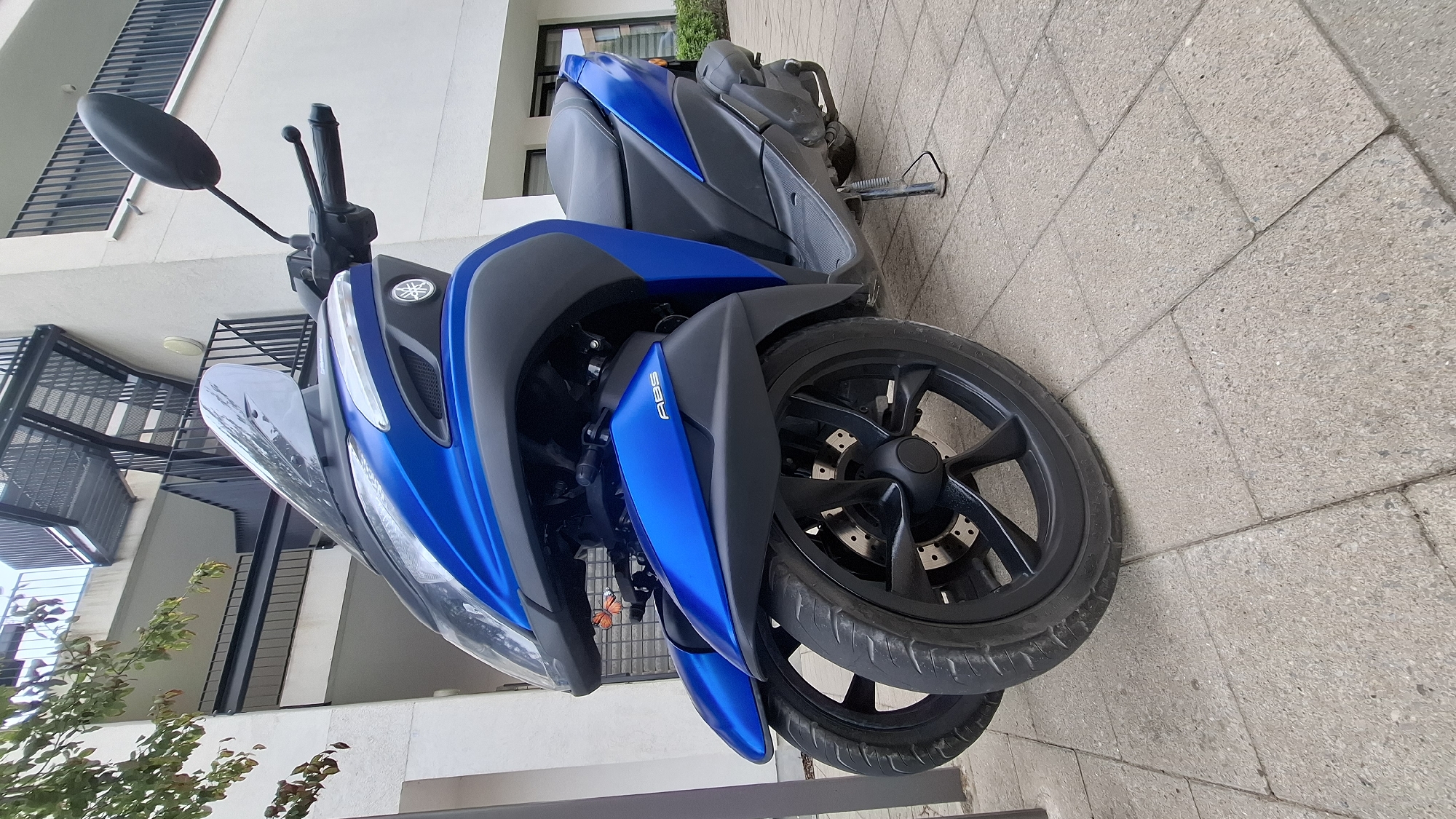 A blue Yamaha Tricity three-wheeled scooter is parked on a concrete paver sidewalk. The scooter has two wheels in the front and one in the rear. The handlebars are black, and the mirrors are also black. The scooter has a windshield and a black seat. A black kickstand is deployed. In the background, a modern building with black railings and stairs is visible. A green tree with some leaves is on the left side of the image. The overall scene is outdoors in an urban or suburban environment.