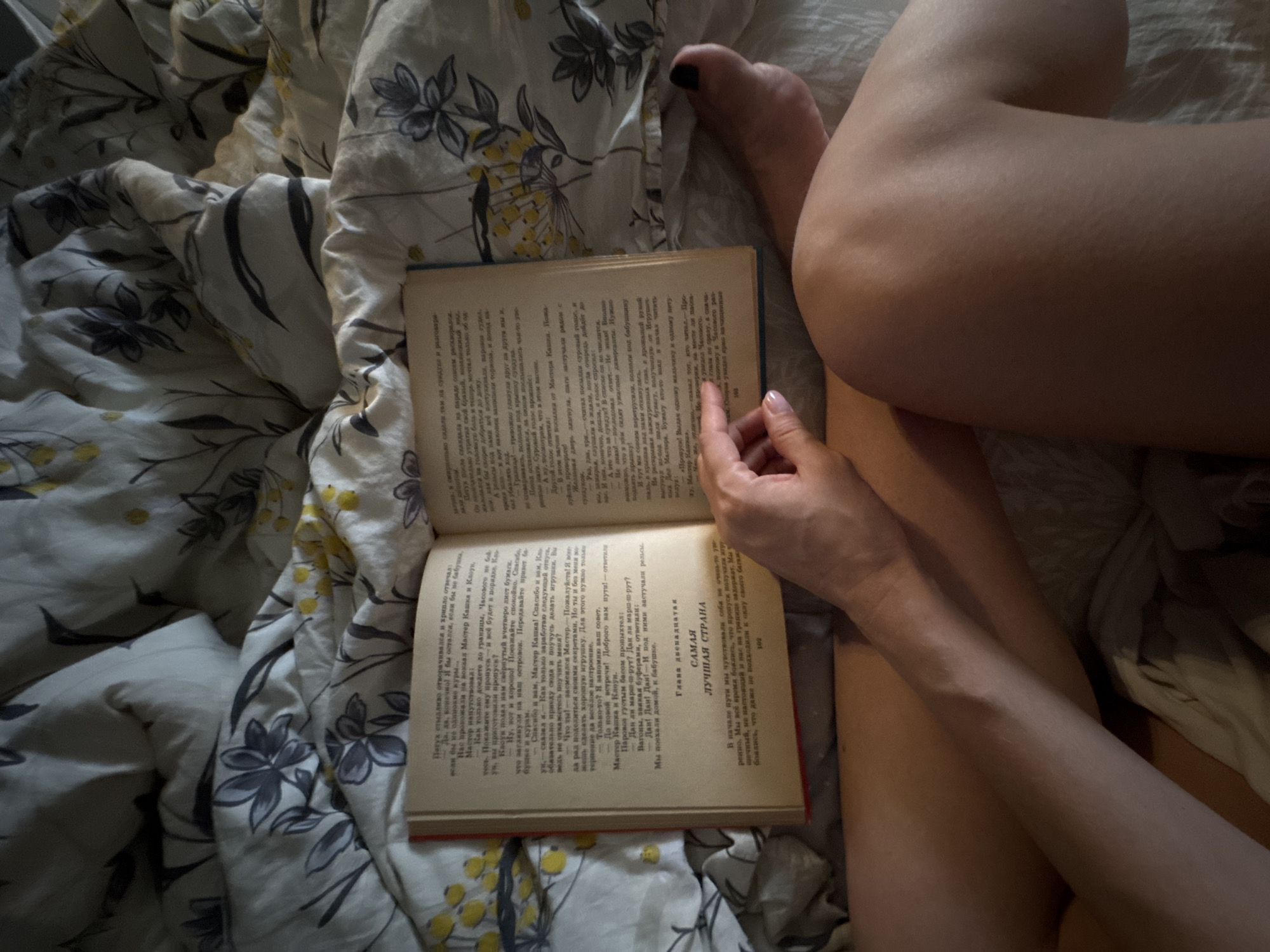 A close-up, high-angle shot captures a person's legs and a book opened on a bed with floral bedding. The person's left arm is visible, resting on their left leg, with their hand holding the book open to a page with Russian text. Their right foot is also visible, with black nail polish on the toenails. The bedding has a pattern of dark grey flowers and yellow buds. The scene appears to be indoors, likely in a bedroom. There is no identifiable information about the location in Chaikovsky, Russia.