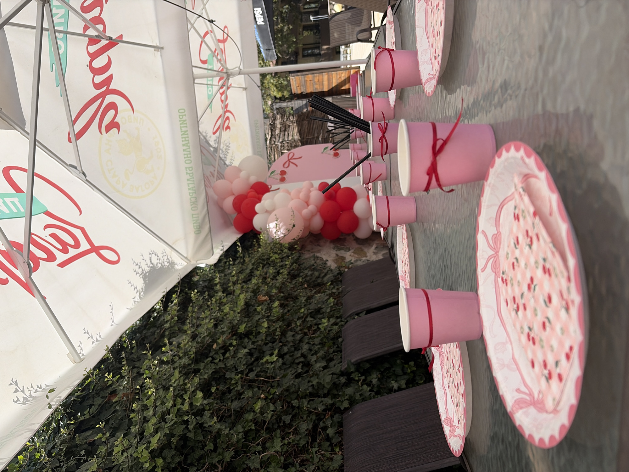 This is an outdoor scene, likely a backyard or patio in Kolarovo, Bulgaria, set for a celebration. A glass-topped table is laden with pink paper cups, each adorned with a red ribbon, and matching paper plates featuring a cherry pattern. Several black straws are gathered in a pink cup. In the background, a large white umbrella, branded with red text and a circular logo, provides shade. To the left, a dense green ivy-covered wall forms a backdrop. To the right, a balloon arch in shades of pink, red, and white stands against a stone wall. Further in the background, wooden structures and trees are visible, along with a distant Pepsi umbrella. The overall aesthetic suggests a children's party or a festive gathering with a pink and cherry theme. There are no people visible in the image.