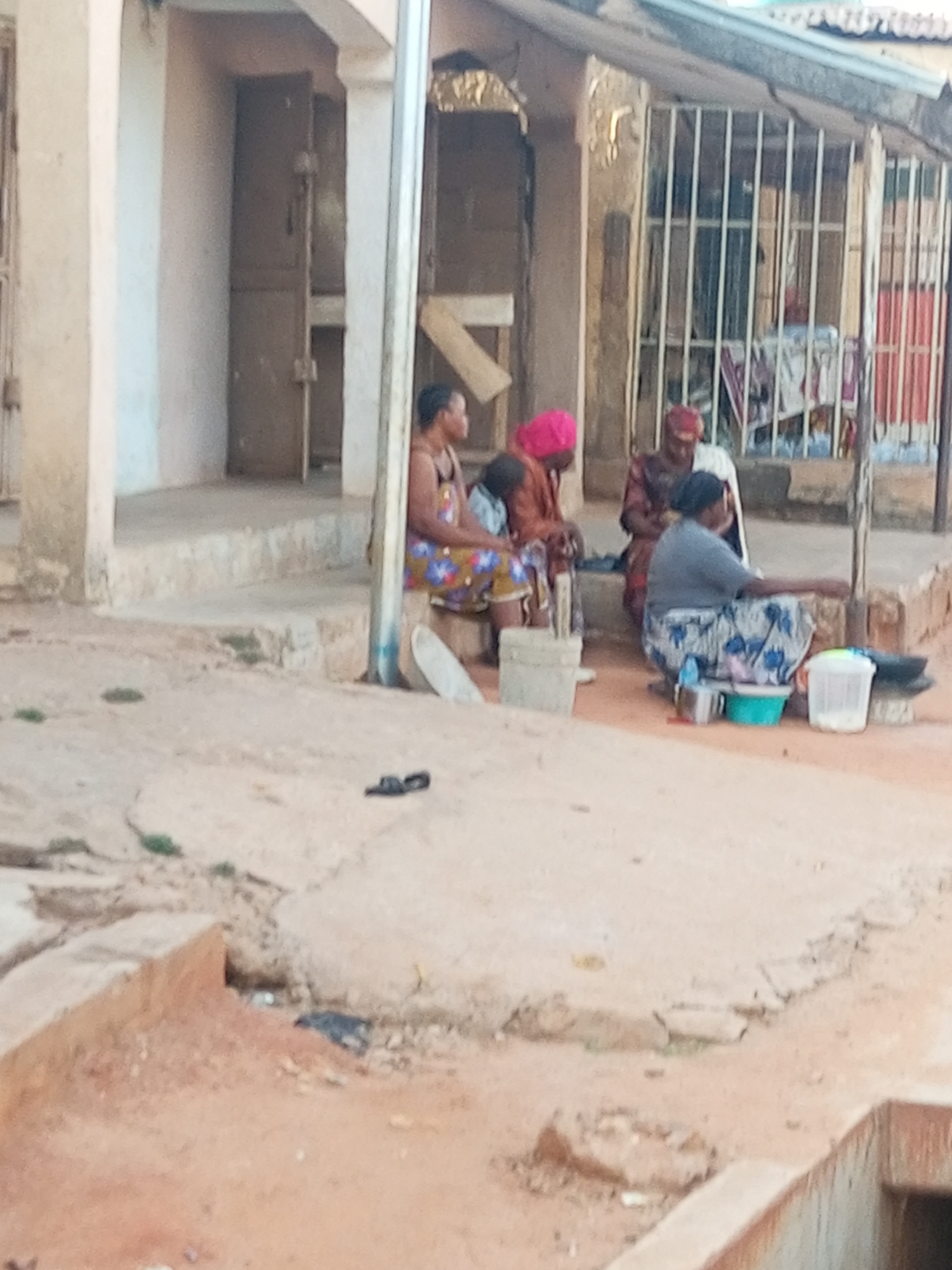 Four women are gathered outside a building in Tempure, Nigeria. One woman is seated and appears to be cooking or preparing food with bowls and utensils. The other three women are seated, possibly observing or participating in the activity. The setting appears to be a residential or communal area with worn surfaces and a partially visible building facade with barred windows. A pair of slippers are seen on the ground.
