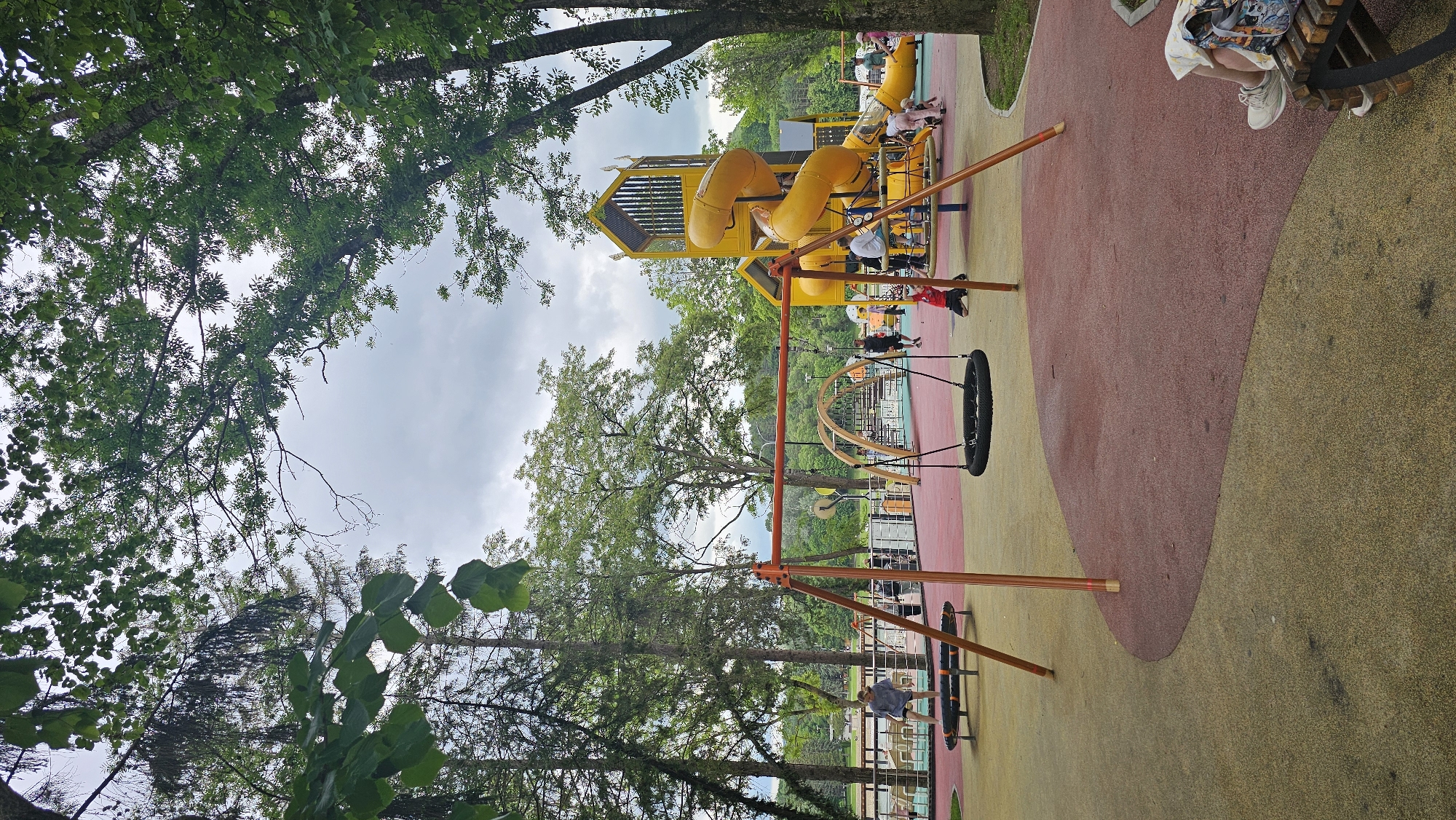 The image depicts a vibrant outdoor playground located in Alikonovka, Russia. The playground features a large yellow multi-level structure with slides and climbing elements, a prominent orange swing set with a black tire swing, and a spinning disc play structure. The ground is covered with a soft, shock-absorbent material in contrasting colors of red and yellow. Several people, including children and adults, are visible enjoying the playground. In the background, tall trees frame the scene, and a body of water, possibly a lake or river, can be seen beyond the playground. The sky is overcast with clouds, suggesting a mild or slightly humid day. The overall impression is of a well-maintained and active recreational space within a natural setting.