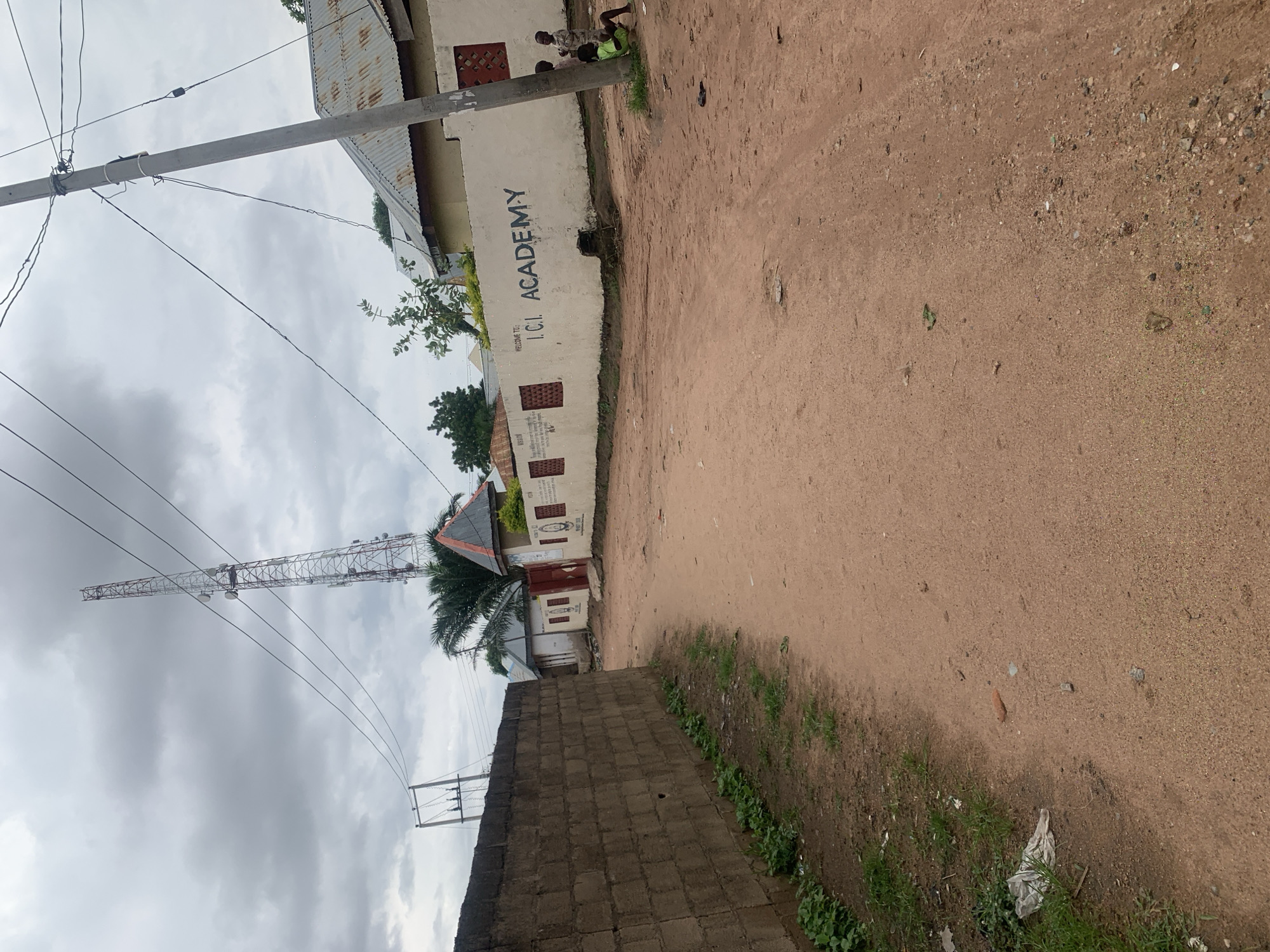 The image depicts a dirt road in Jalingo, Nigeria, leading towards a building with "I.C.I. ACADEMY" written on its wall. A tall telecommunications tower is visible in the background, along with power lines and a utility pole. To the left of the road, a low brick wall runs alongside the path. Several children are present near the academy building, with two sitting by the utility pole and one standing further away. The sky is overcast with clouds. The environment appears to be a semi-urban or rural area within Jalingo.