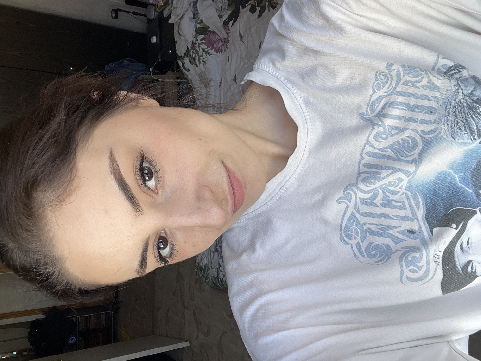 A young woman with brown hair and eyes is facing the camera in a close-up shot. She is wearing a white, long-sleeved shirt with a blue design. The background includes a floral bedspread and a glimpse of what appears to be a room. The location in the city of Moscow, Russia, is not discernible from the provided visual information.