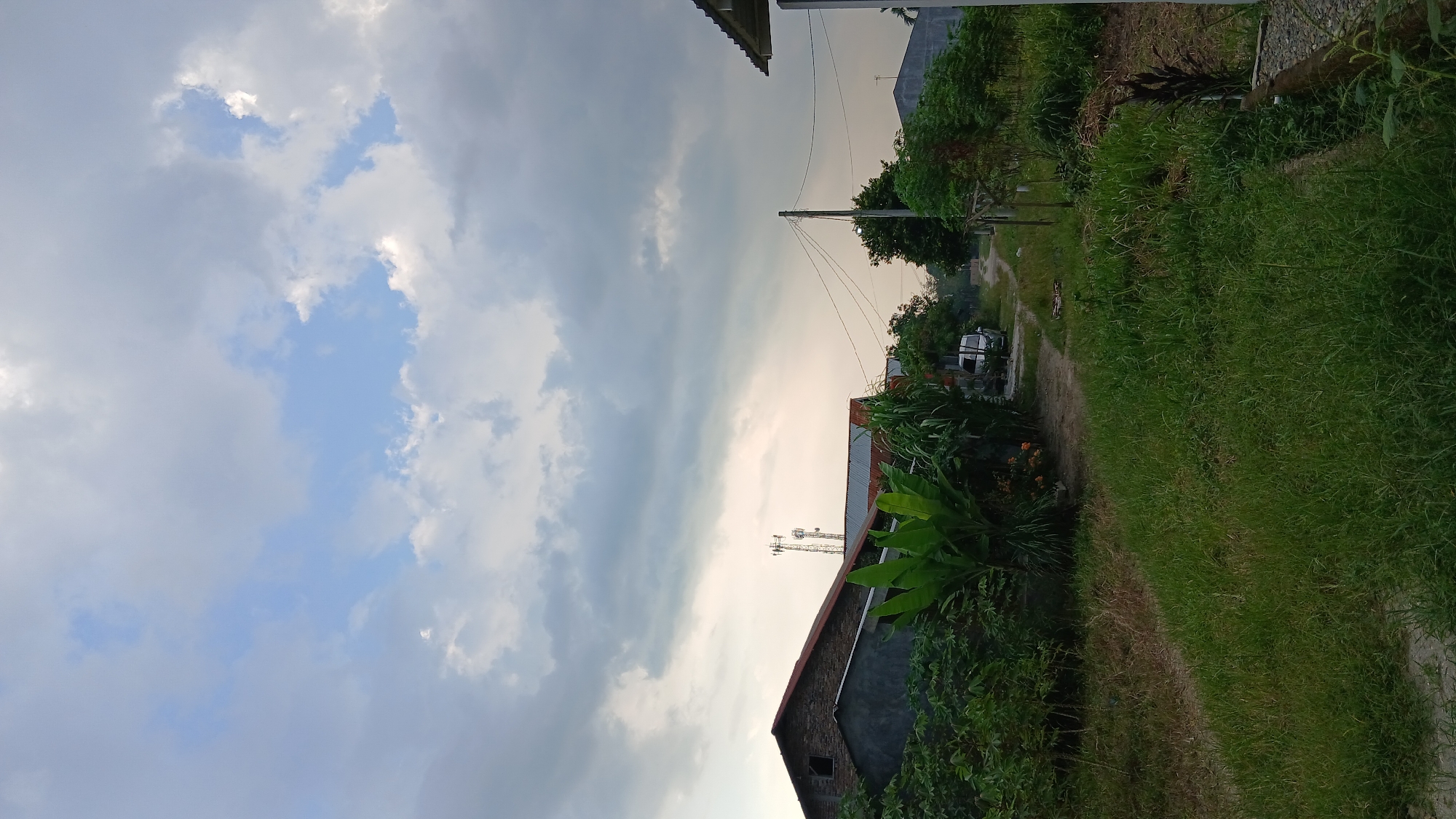 The image depicts a rural scene in Klambir Lima Kebun, Indonesia, under a cloudy sky. In the foreground, a path covered with lush green grass leads into the distance. To the left, there are buildings with dark roofs and concrete walls, surrounded by dense green foliage, including banana plants. Further into the background, a dirt road is visible, with a white car parked on the right side. Two cellular towers stand tall against the sky, and a utility pole with power lines is also present. The overall impression is one of a residential area on the outskirts of a town, with a mix of man-made structures and abundant vegetation.